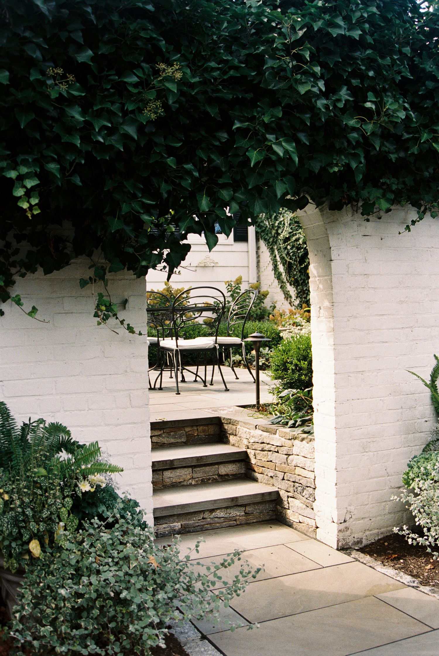 Garden patio area with white painted walls and built-in stone benches surrounded by lush greenery.
