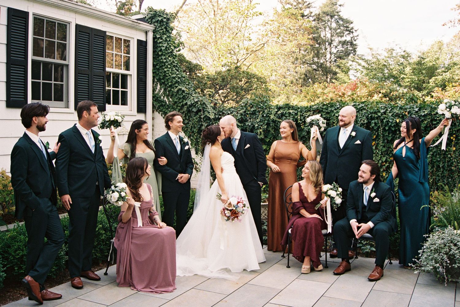 Outdoor wedding ceremony with guests seated in white chairs against dark green building.