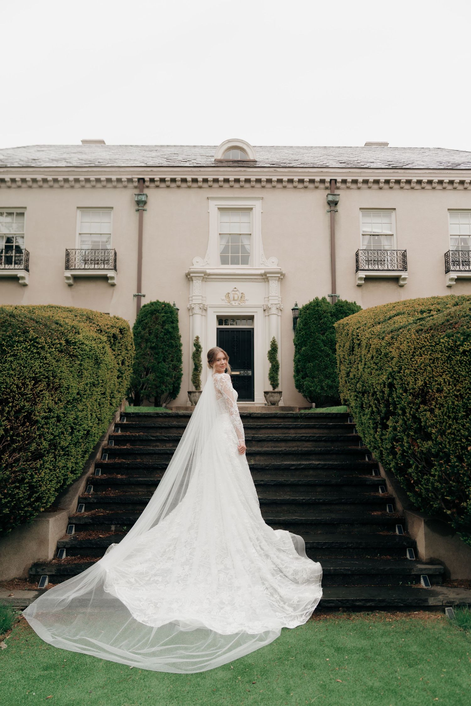 Bride in stunning lace wedding dress with dramatic cathedral train standing before grand estate staircase.