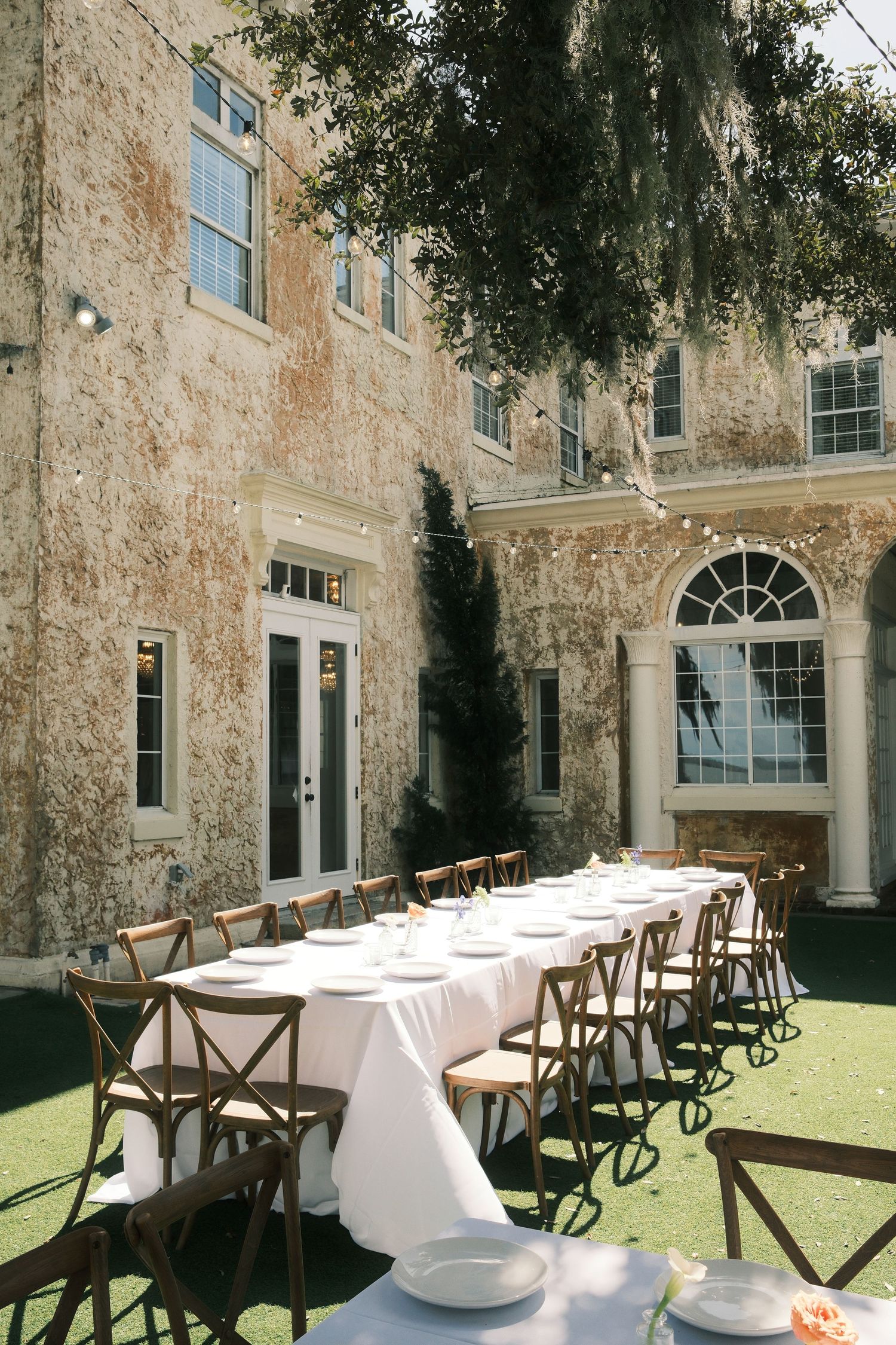 Elegant outdoor reception setup with long white tables and wooden cross-back chairs against a historic stone building.