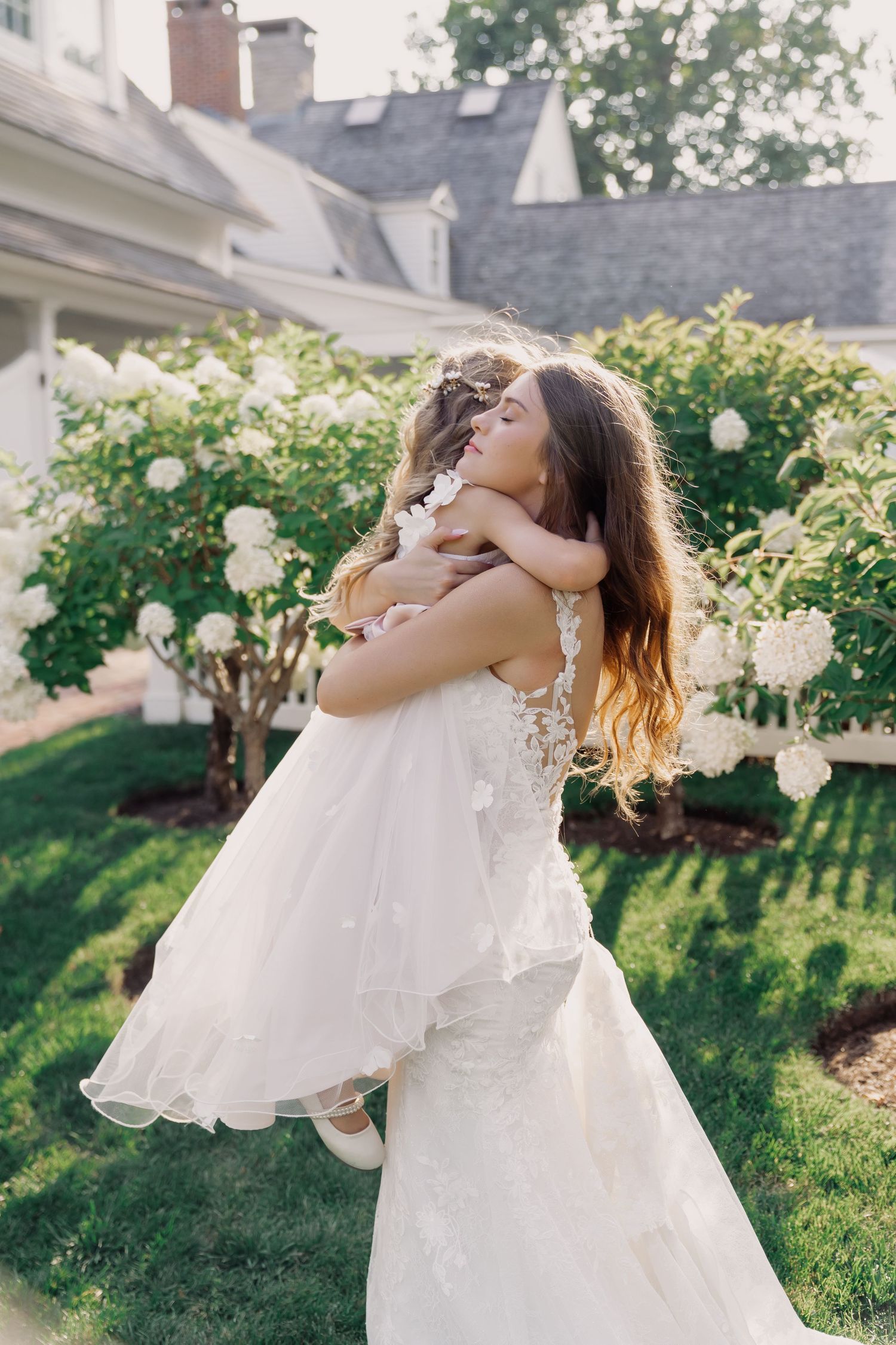 A mother and daughter in white dresses dance together on a sunny lawn with trees and houses in the background.