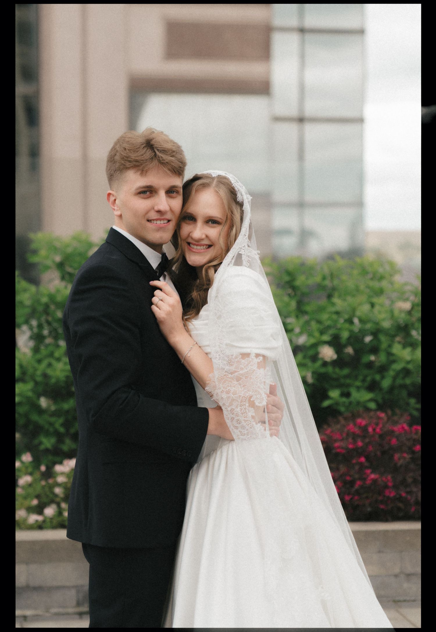 A romantic wedding portrait shows a couple embracing outdoors with a veil and black tuxedo against a garden backdrop.