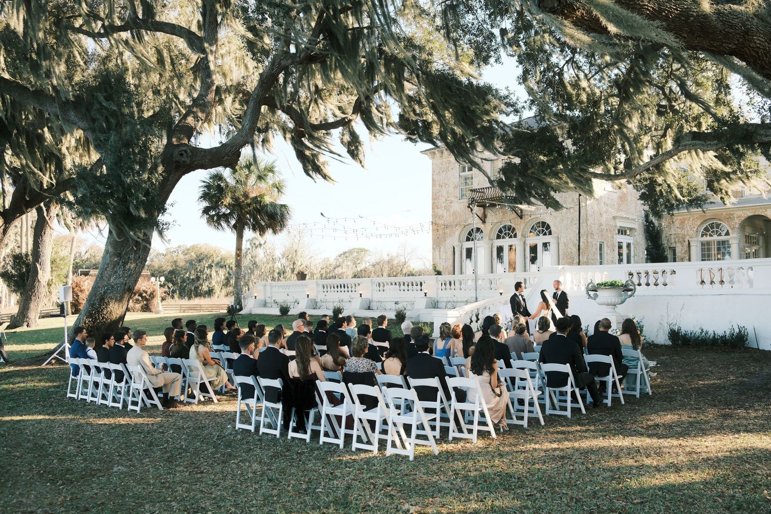 White folding chairs arranged in rows under oak trees at an outdoor wedding venue during golden hour.