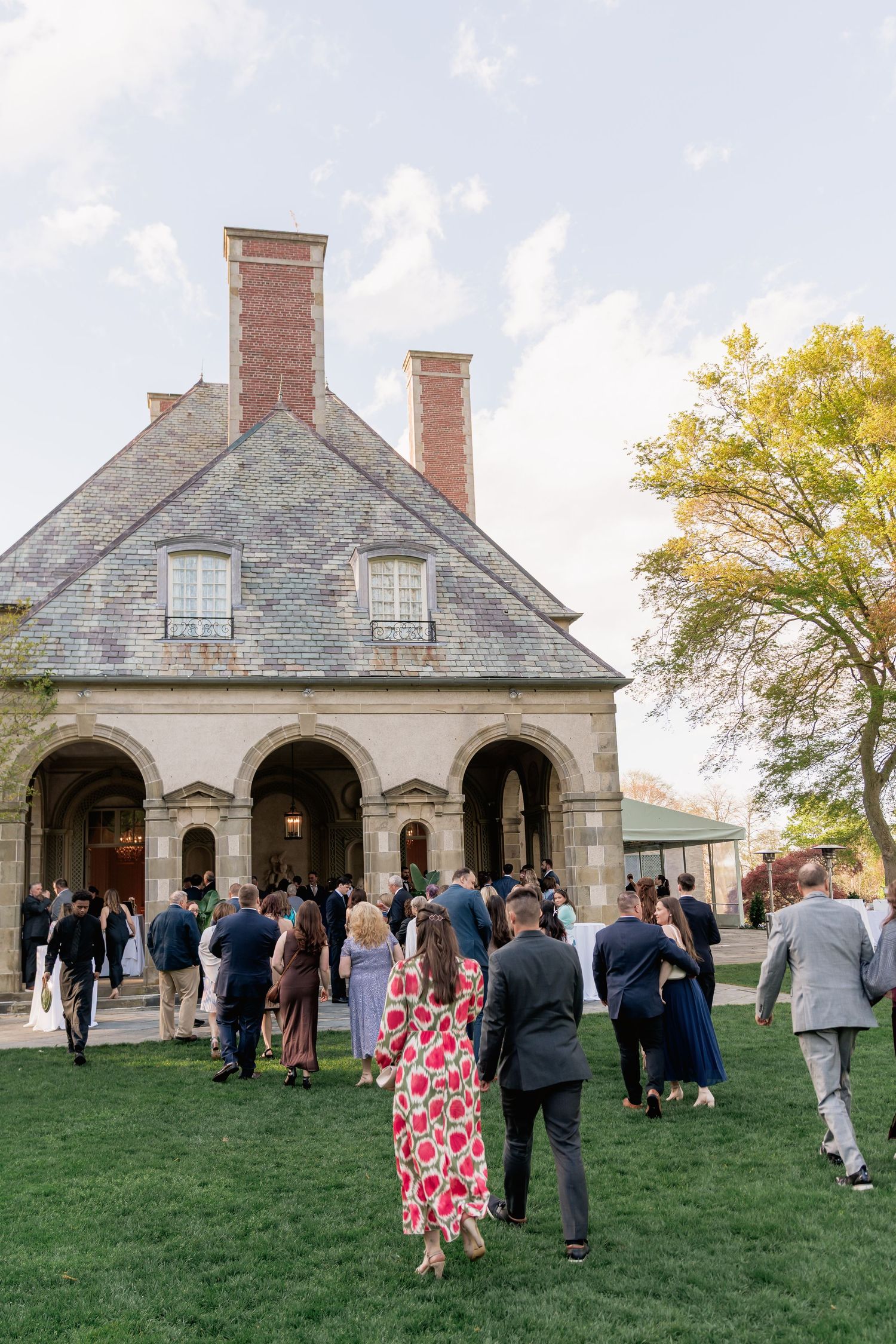 Guests gather on green lawn outside historic stone building with arched portico and brick chimneys at elegant outdoor event.