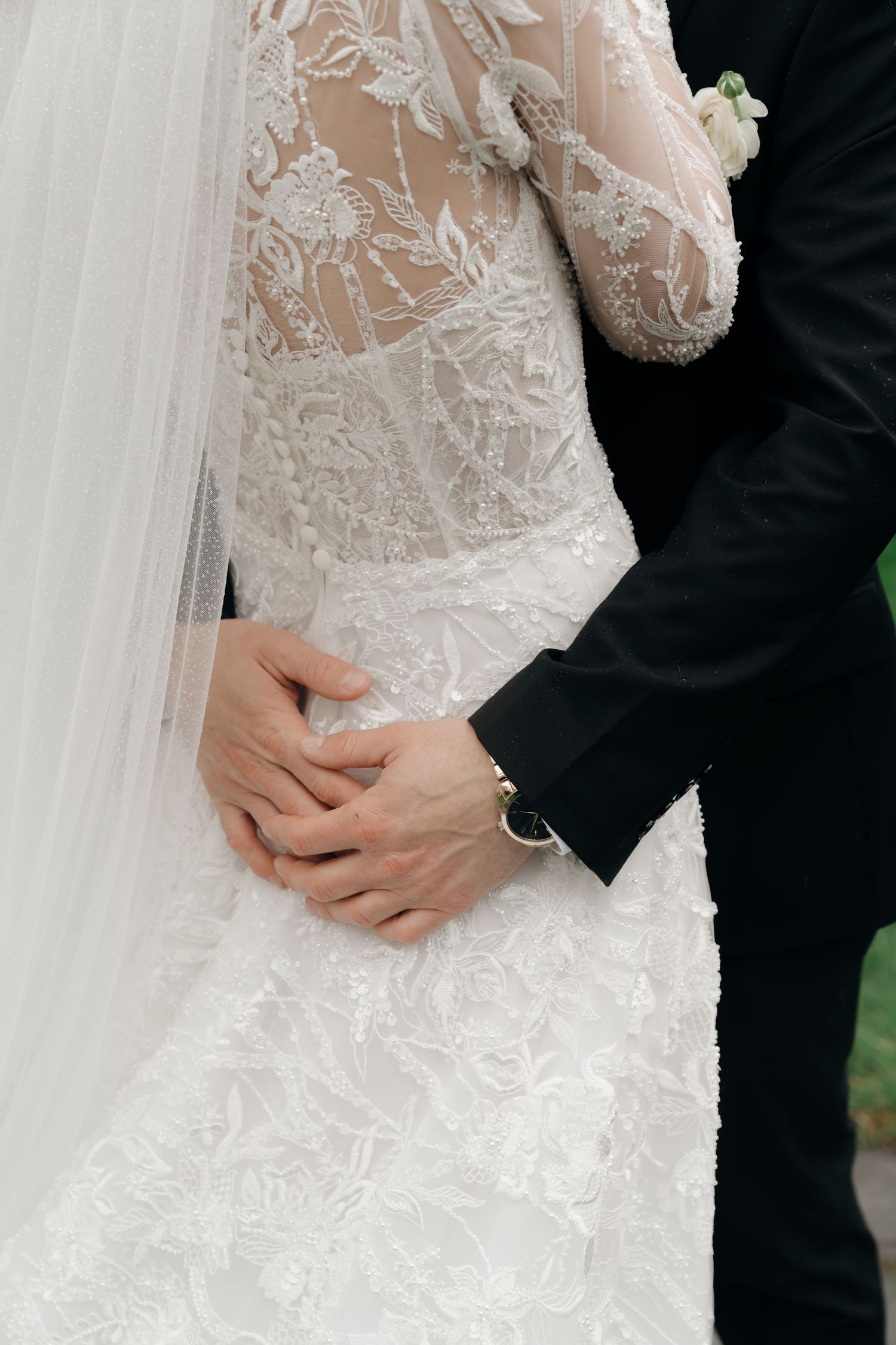 Bride and groom holding hands, showcasing intricate lace wedding dress details and groom's black suit sleeve.