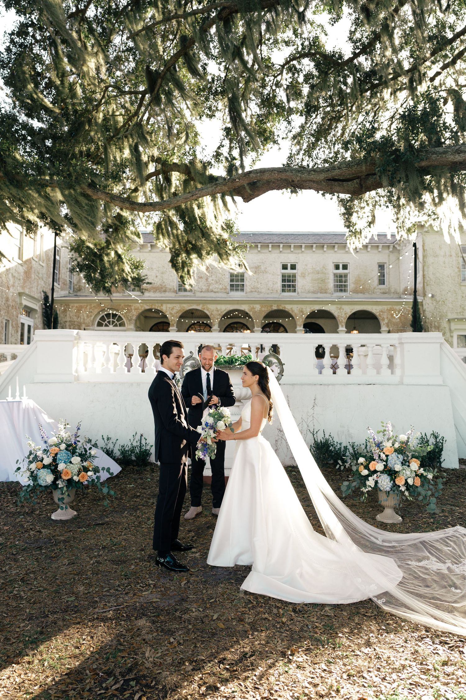 Wedding ceremony setup in a beautiful courtyard with white chairs and floral arrangements under Spanish moss draped trees.