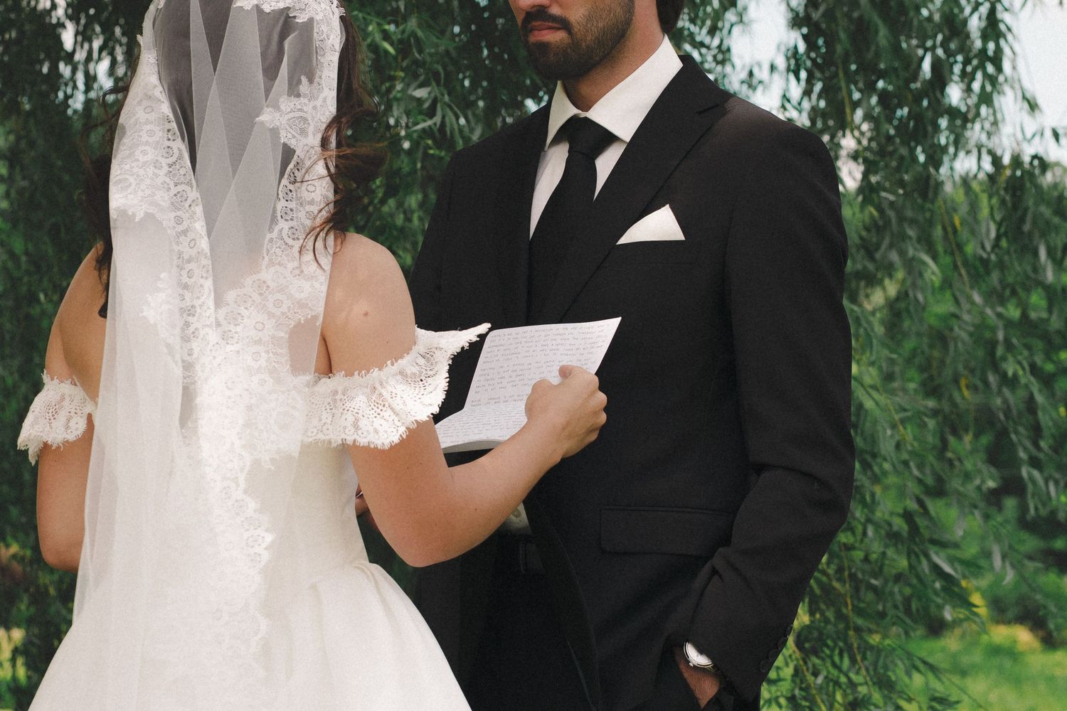 Bride and groom exchanging vows outdoors, bride in lace wedding dress with veil, groom in black suit with tie.