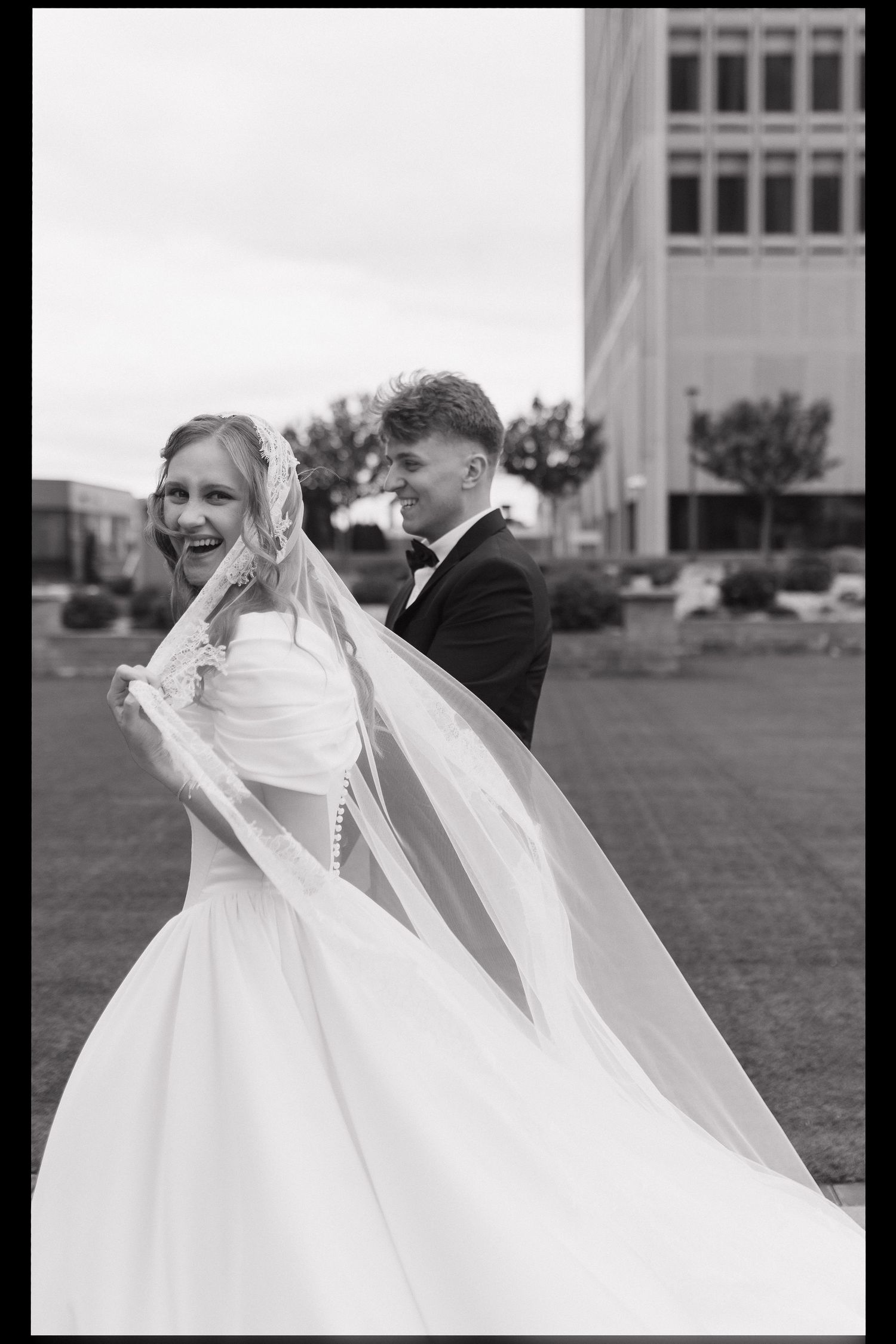 A black and white wedding photo showing a bride in a flowing white gown with her veil caught in the wind.