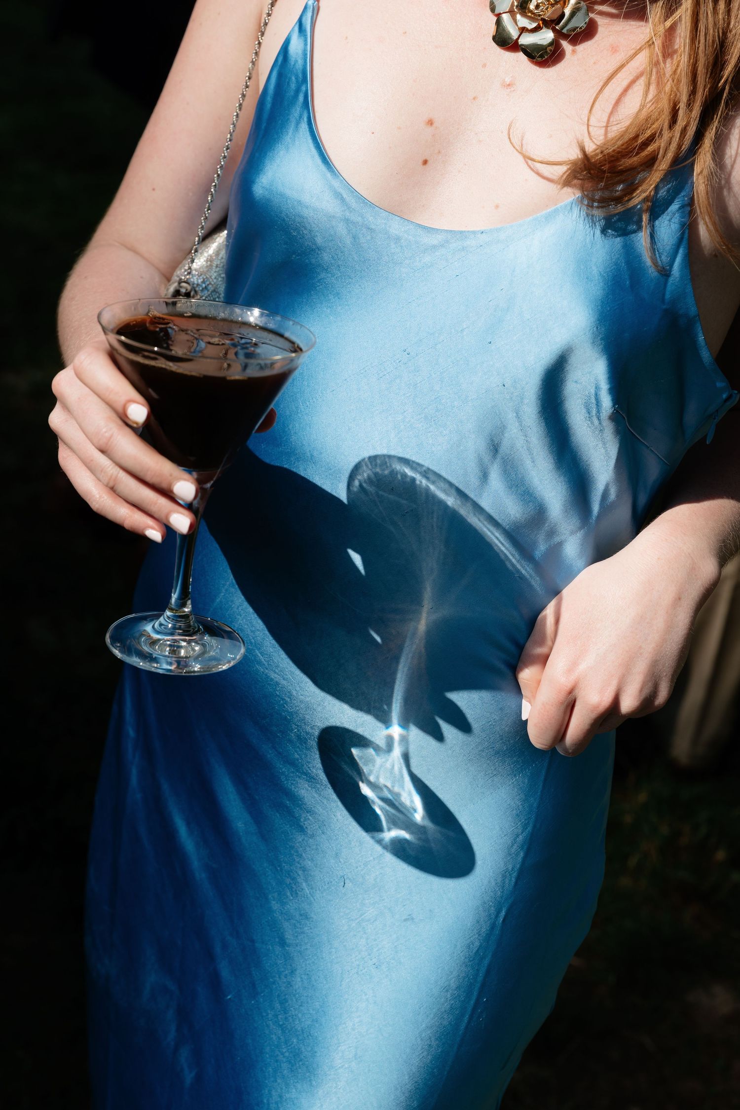 Woman in blue satin slip dress holding a red wine glass with sunlight casting elegant shadow on fabric.