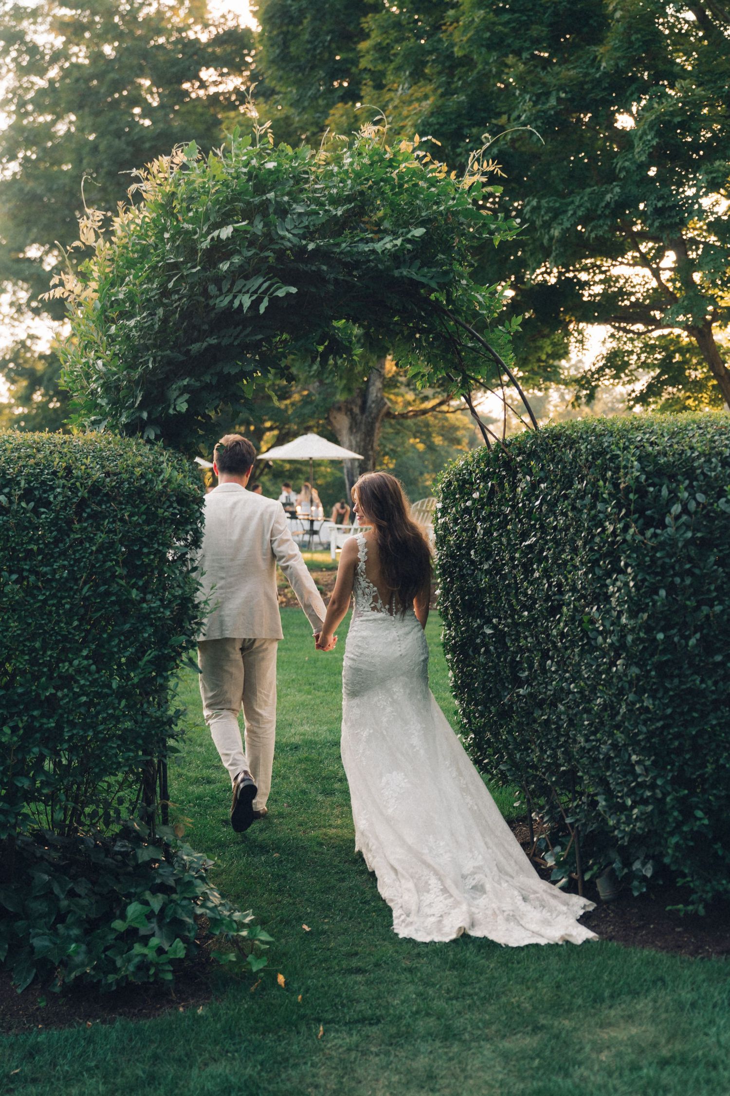 A couple in wedding attire walks along a garden path during sunset with lush green hedges in the background.