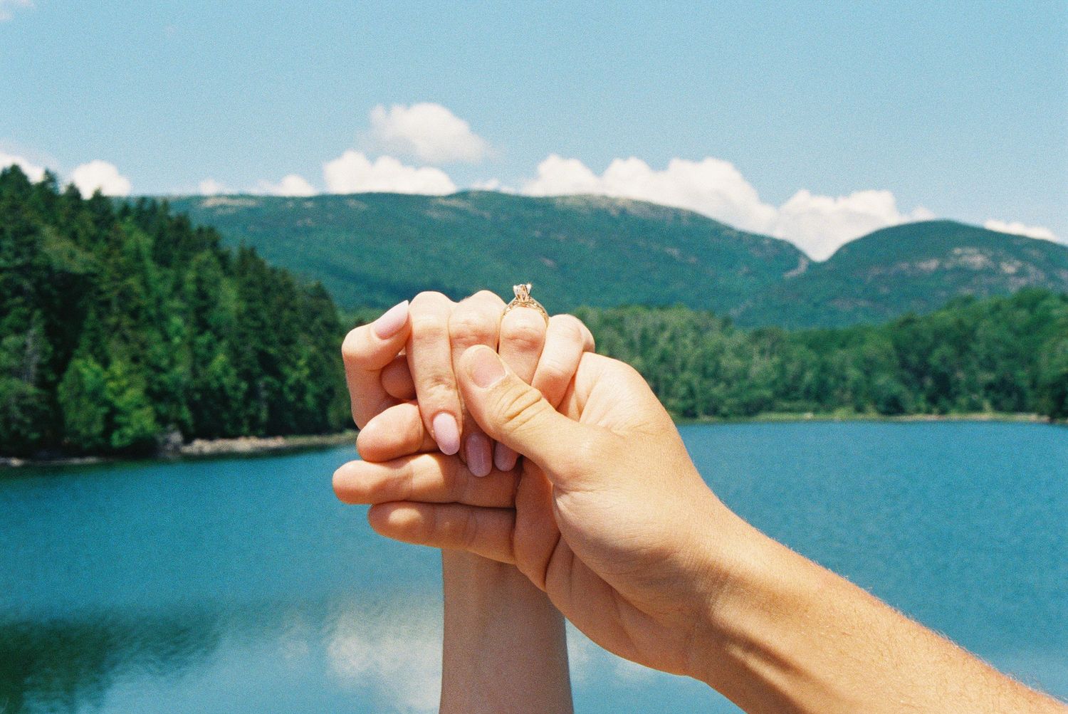 Two hands clasped together near a serene mountain lake surrounded by lush green trees under a blue sky.