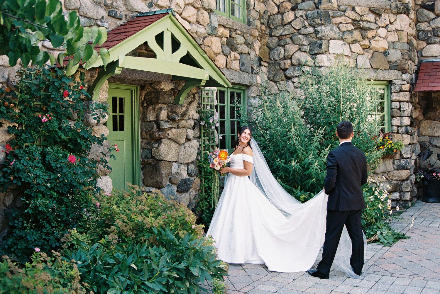 Bride in white gown with veil held by groom near stone building with lush greenery and yellow bouquet.