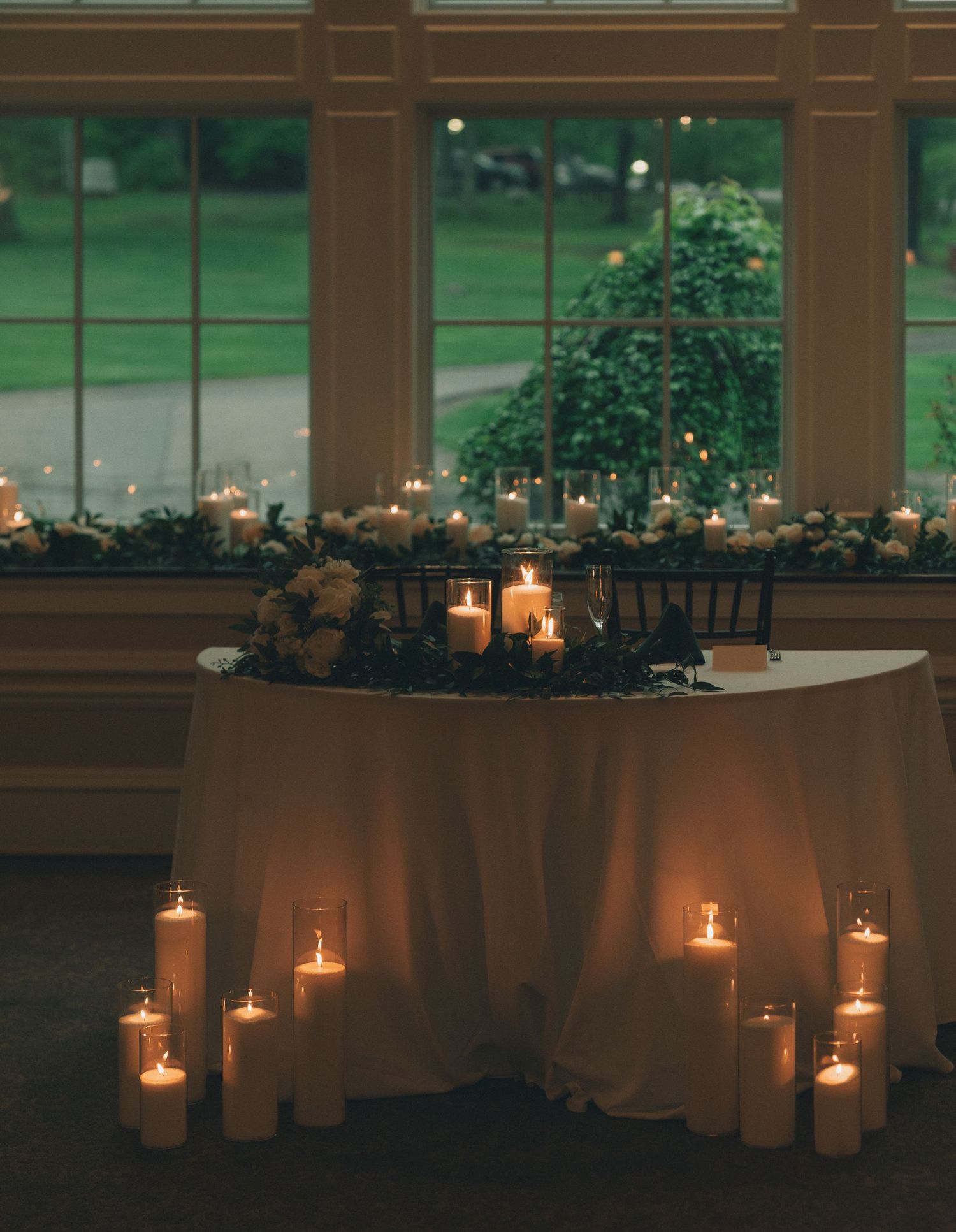 Elegant wedding reception setup with glowing candles and white tablecloths in a windowed venue.