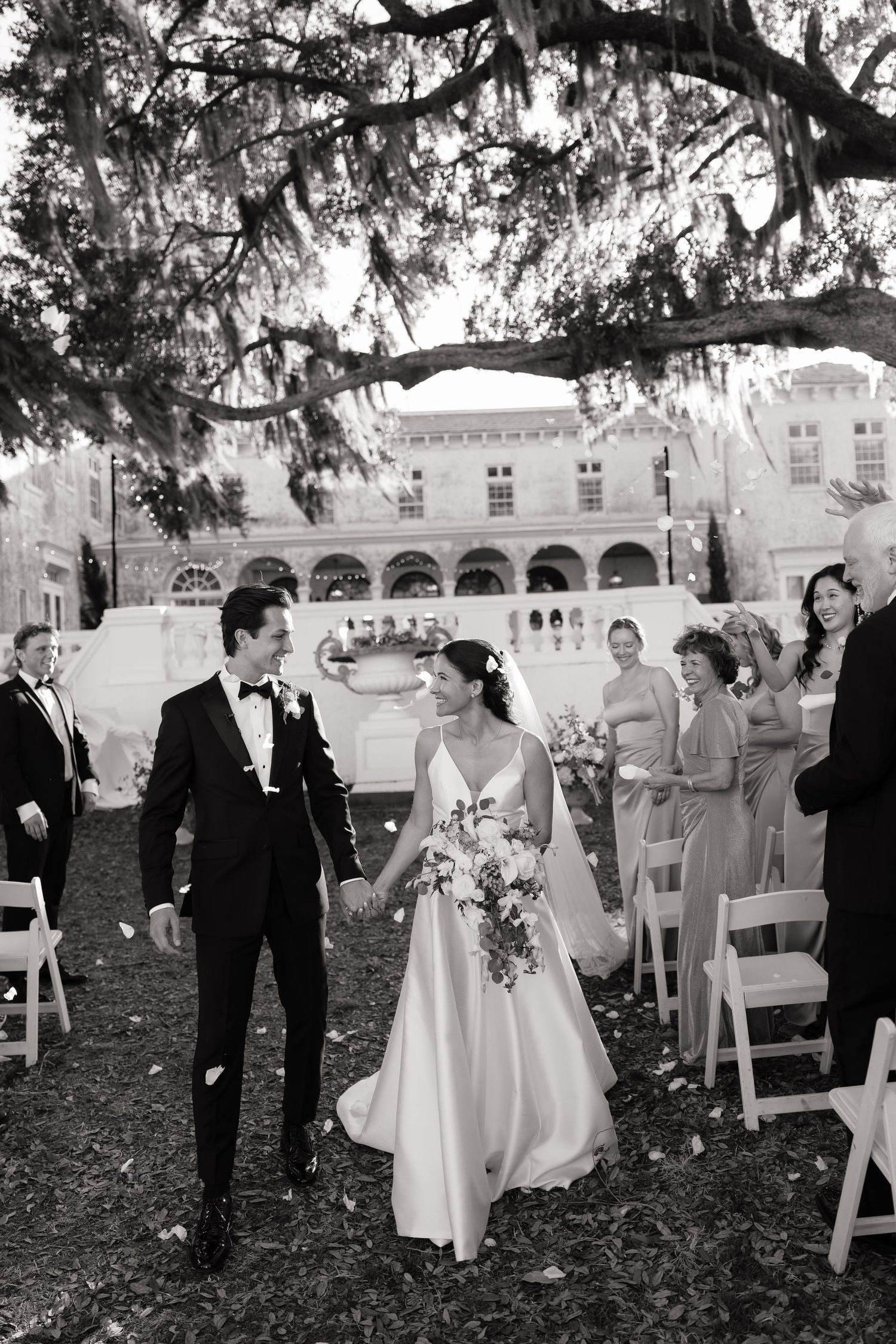 Elegant outdoor wedding reception under oak trees with guests mingling around white chairs.