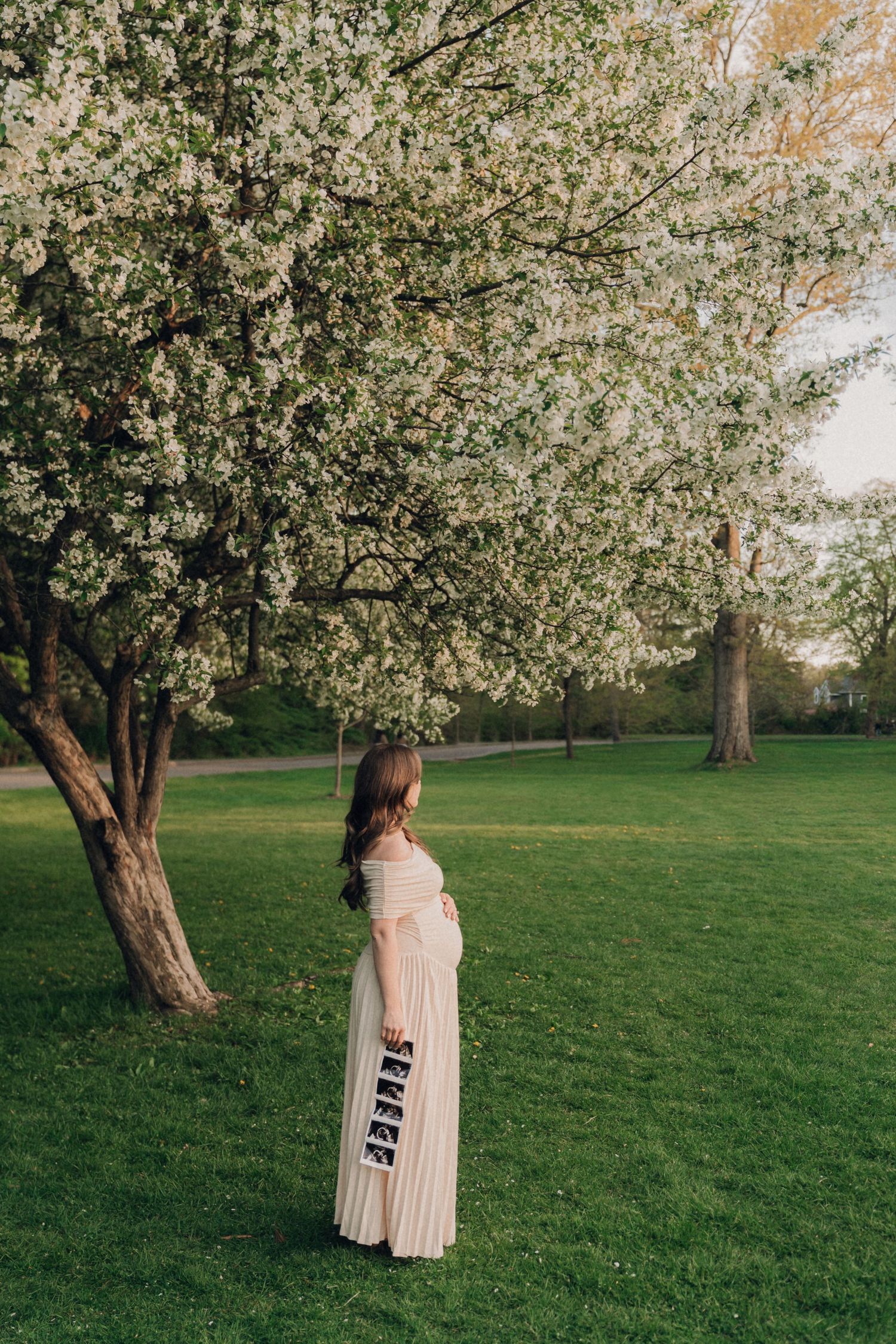 Pregnant woman in a cream maxi dress standing beside a blooming white flowering tree in a lush green park.