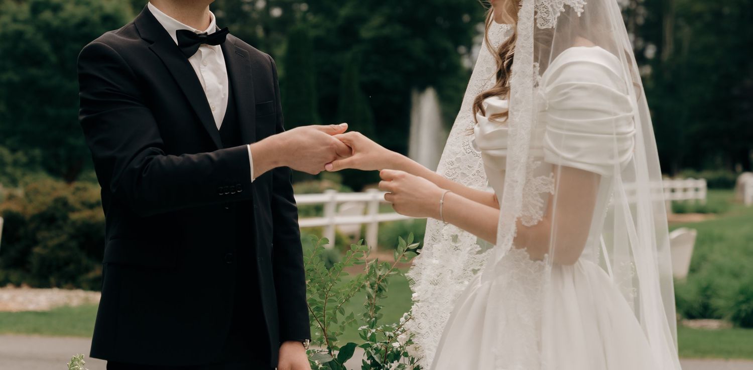 A couple in formal wedding attire stands close together while exchanging drinks outdoors in a garden setting.