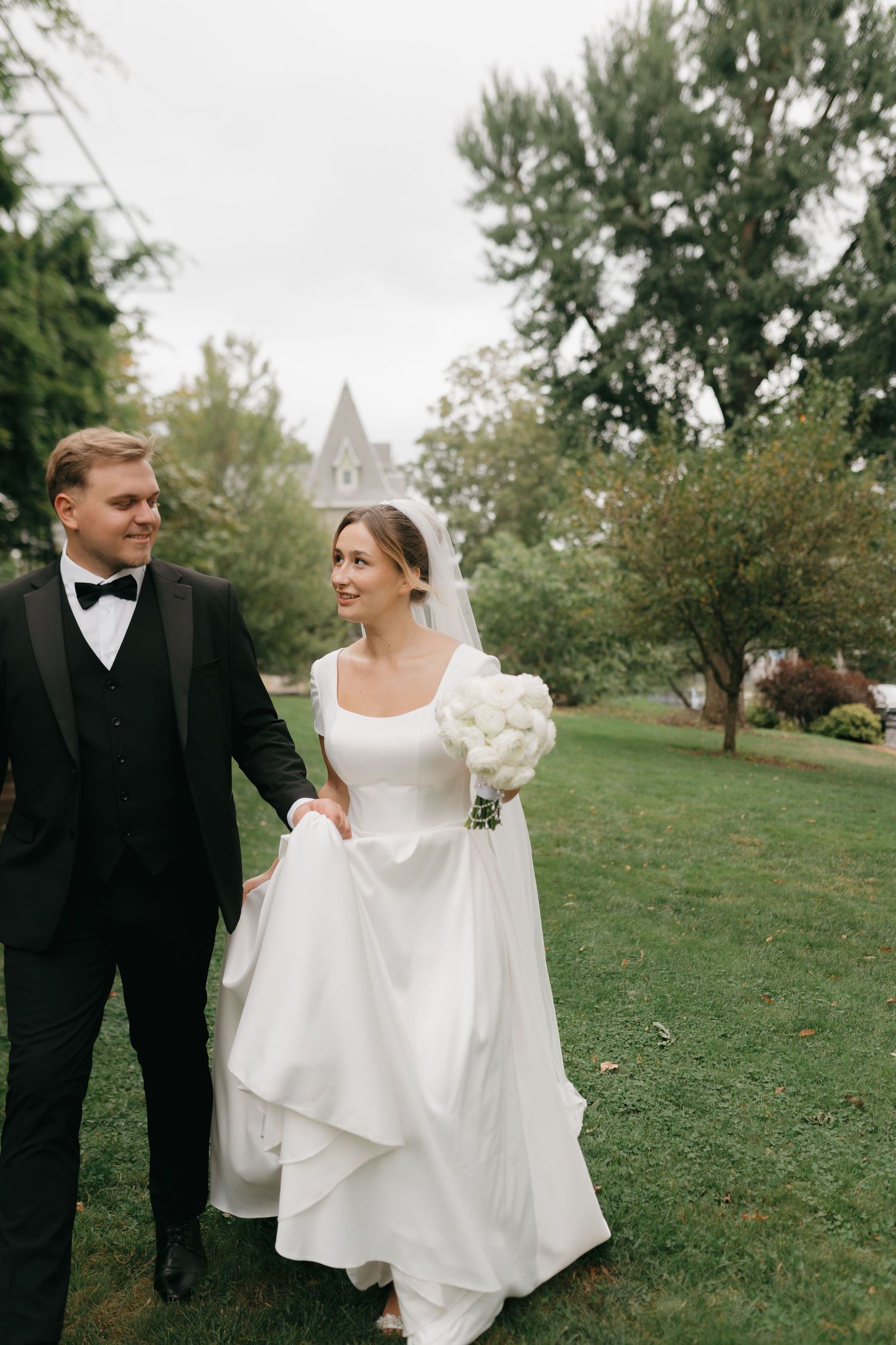 Newlyweds walking together on a lawn with a church spire visible in the background.