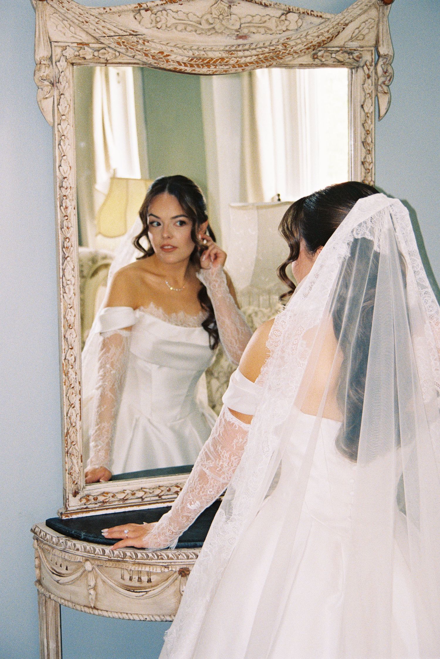 Bride in off-shoulder lace wedding gown and veil adjusts earring while gazing into ornate vintage mirror.