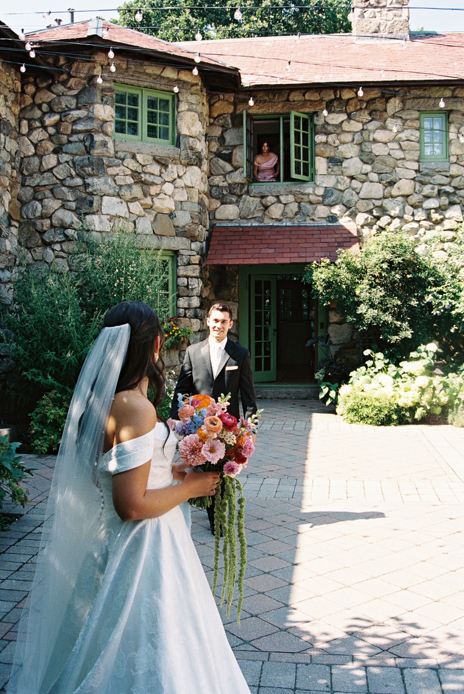 Bride in white gown holds colorful bouquet facing groom outside a stone building during wedding first look.