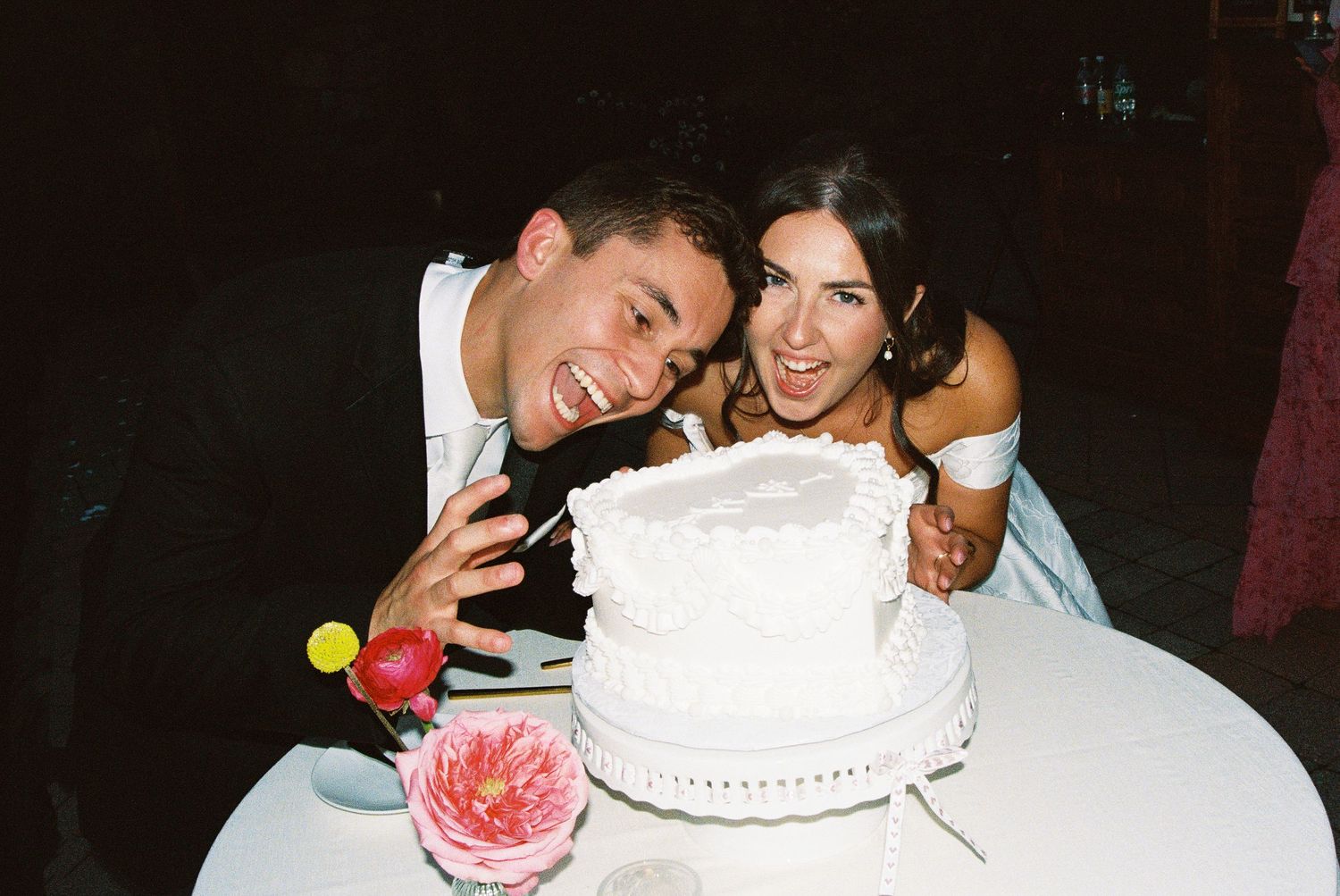 Joyful newlyweds laughing beside a white wedding cake at their reception celebration.