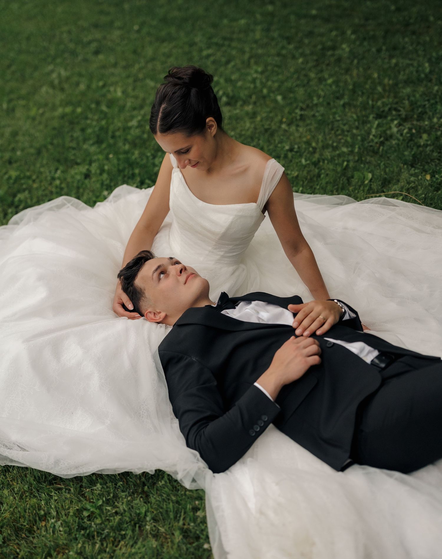 Bride in white gown sits on grass while groom in black tuxedo rests his head in her lap outdoors.
