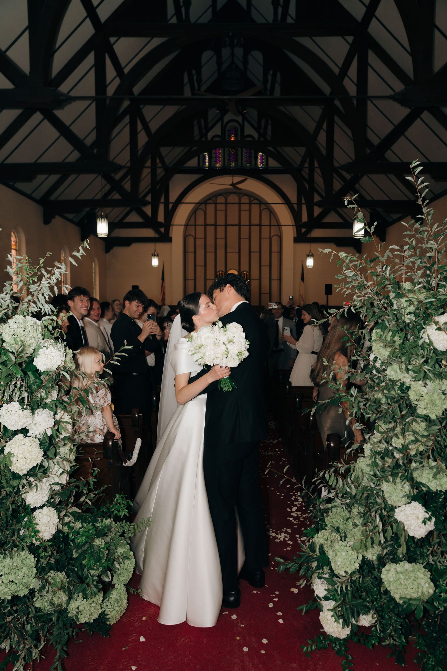 A couple shares a romantic moment under a vaulted chapel ceiling surrounded by lush white floral arrangements.