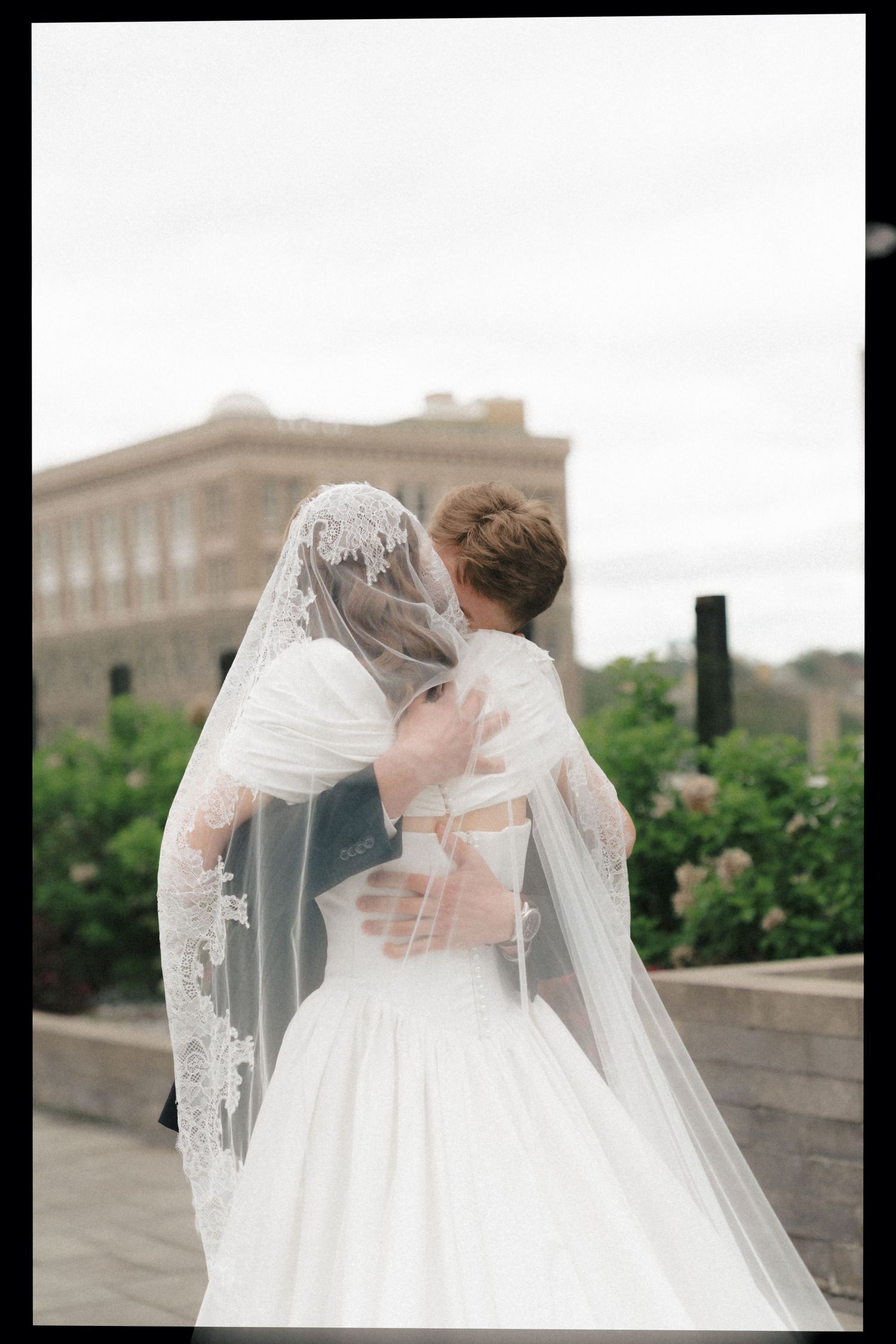 A romantic embrace between newlyweds on their wedding day with a flowing white veil and elegant dress against a stone building.