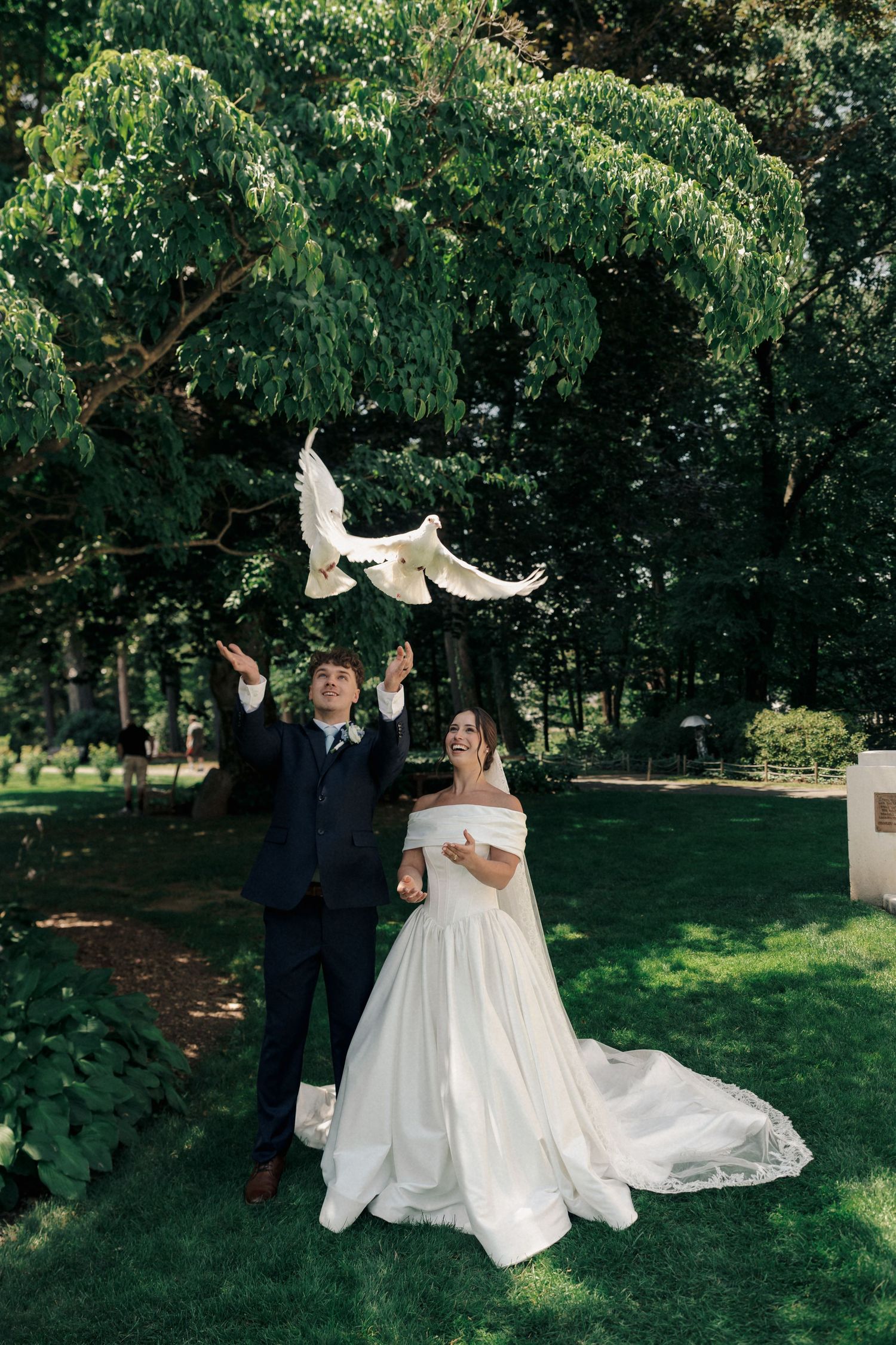 Bride and groom releasing white doves during outdoor wedding ceremony surrounded by lush green trees.