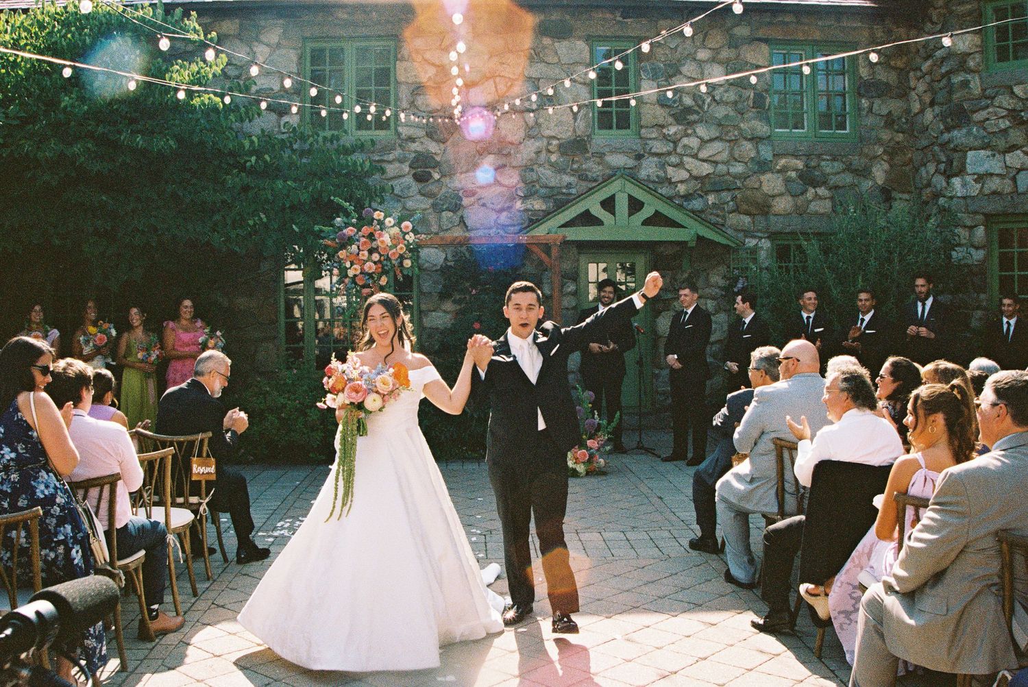 Bride and groom walking down the aisle at an outdoor wedding ceremony with string lights and guests seated alongside.