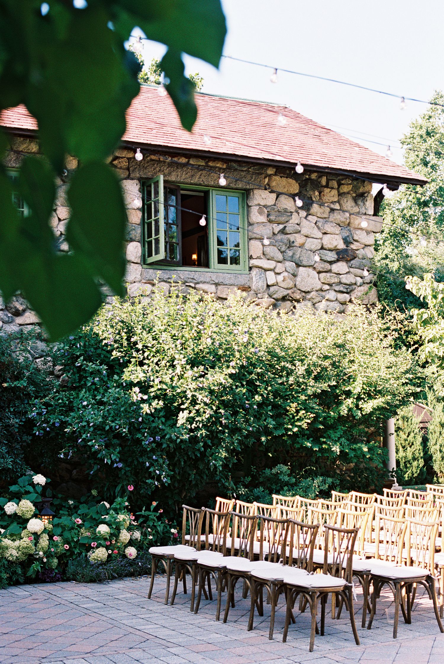 Rows of wooden chairs set on a stone patio for an outdoor wedding ceremony beside a rustic stone building with lush greenery.