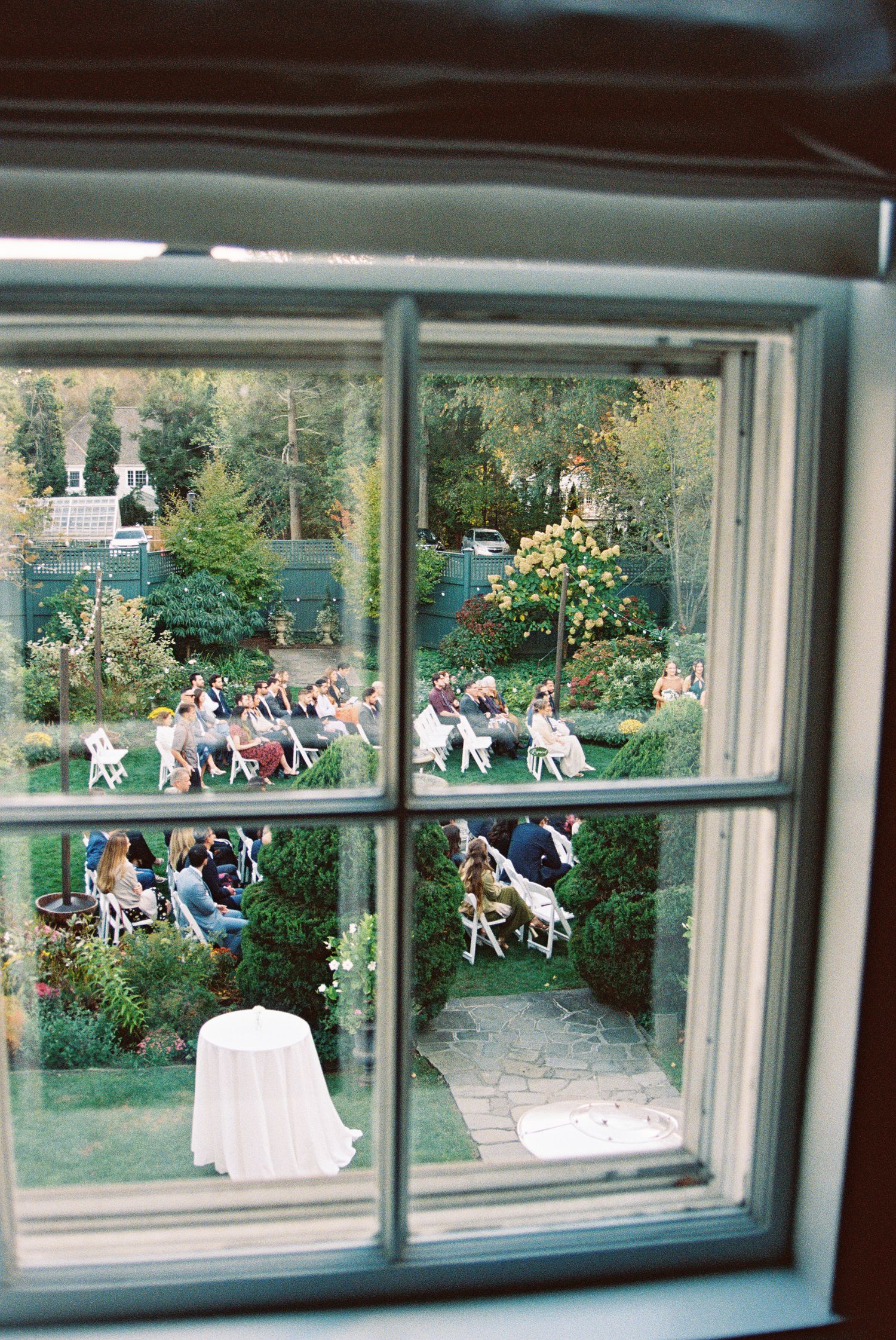 View through a large window showing guests seated in garden chairs for an outdoor ceremony.