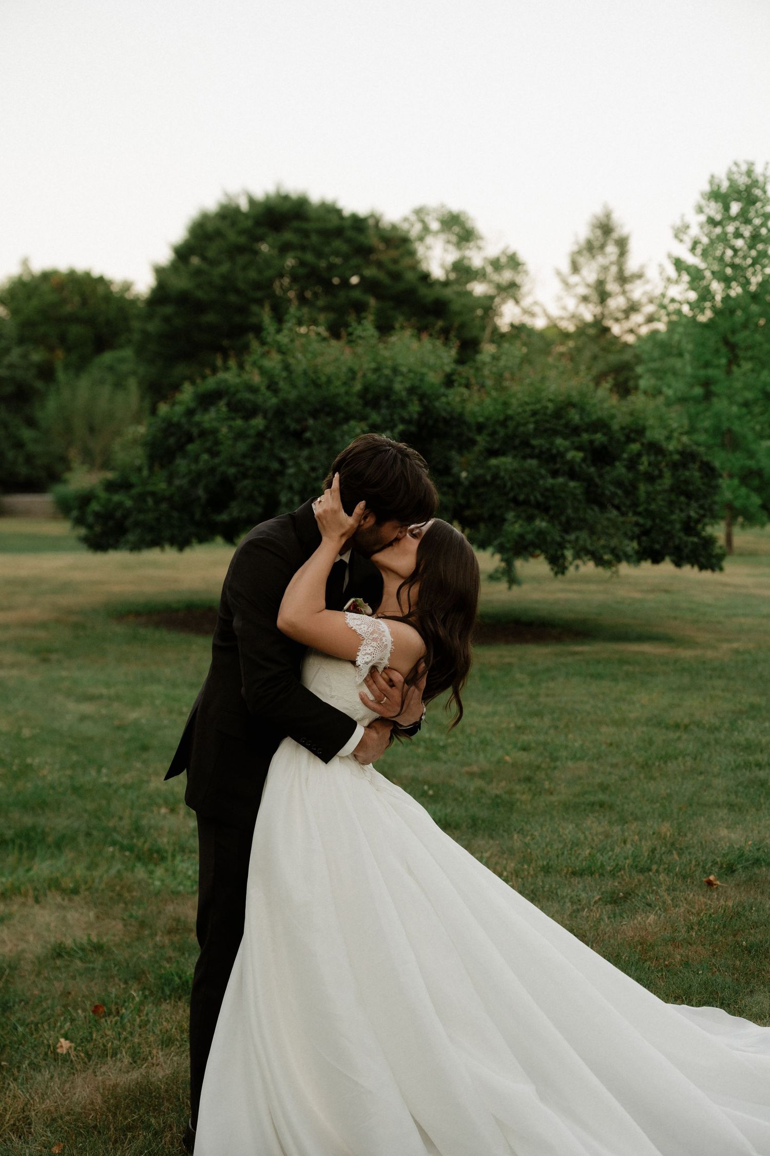 Bride and groom share a romantic kiss in a lush green garden, brides gown trailing elegantly.