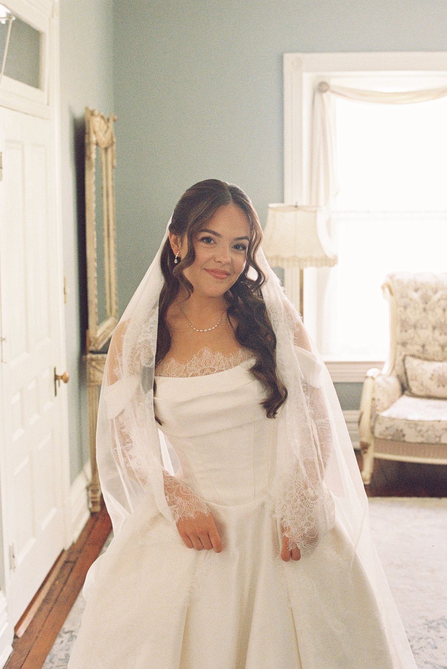 Bride in white ball gown and lace veil smiling in an elegant room with soft natural light.