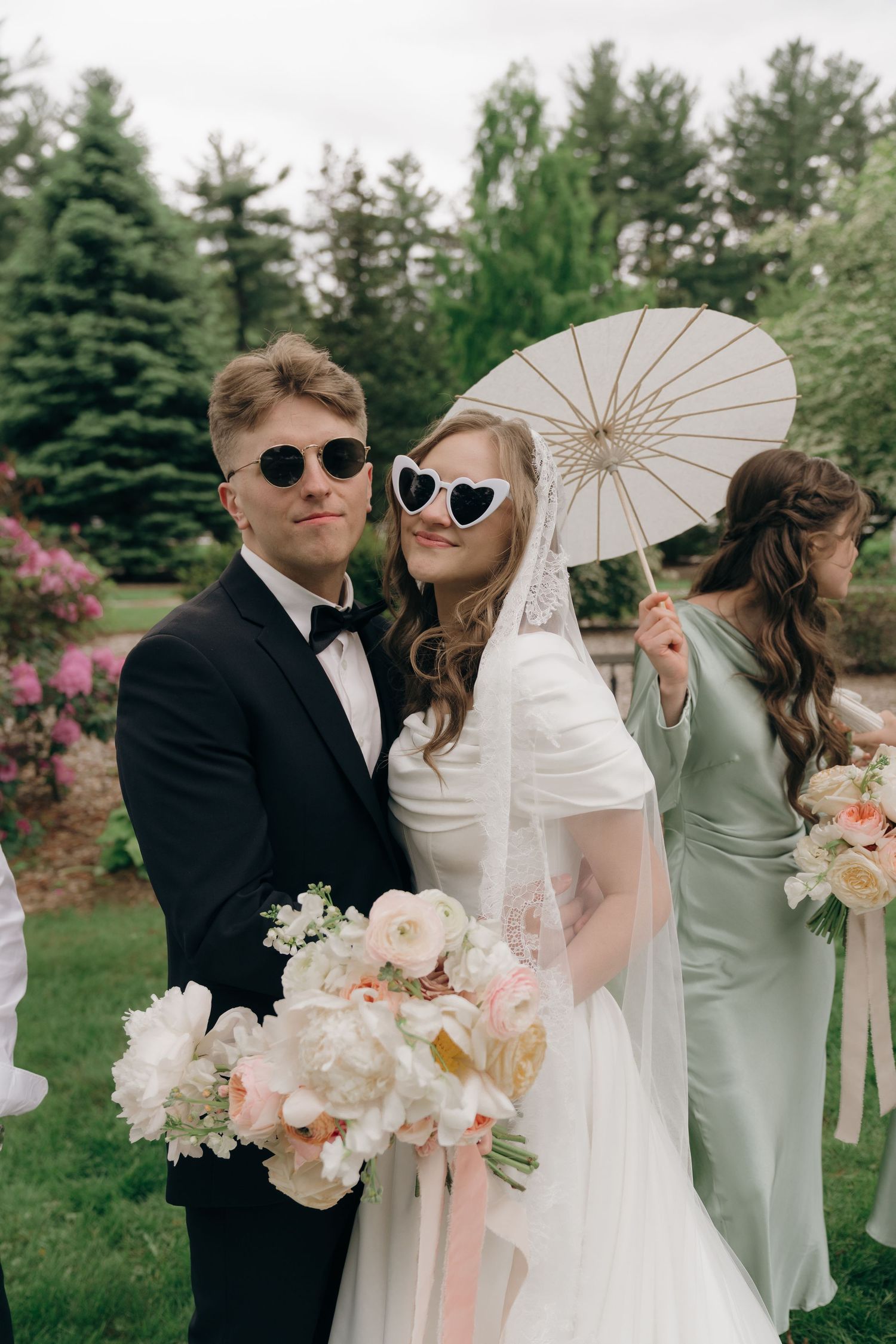 Wedding party poses with parasols and sunglasses in a sunny garden setting with flowers.