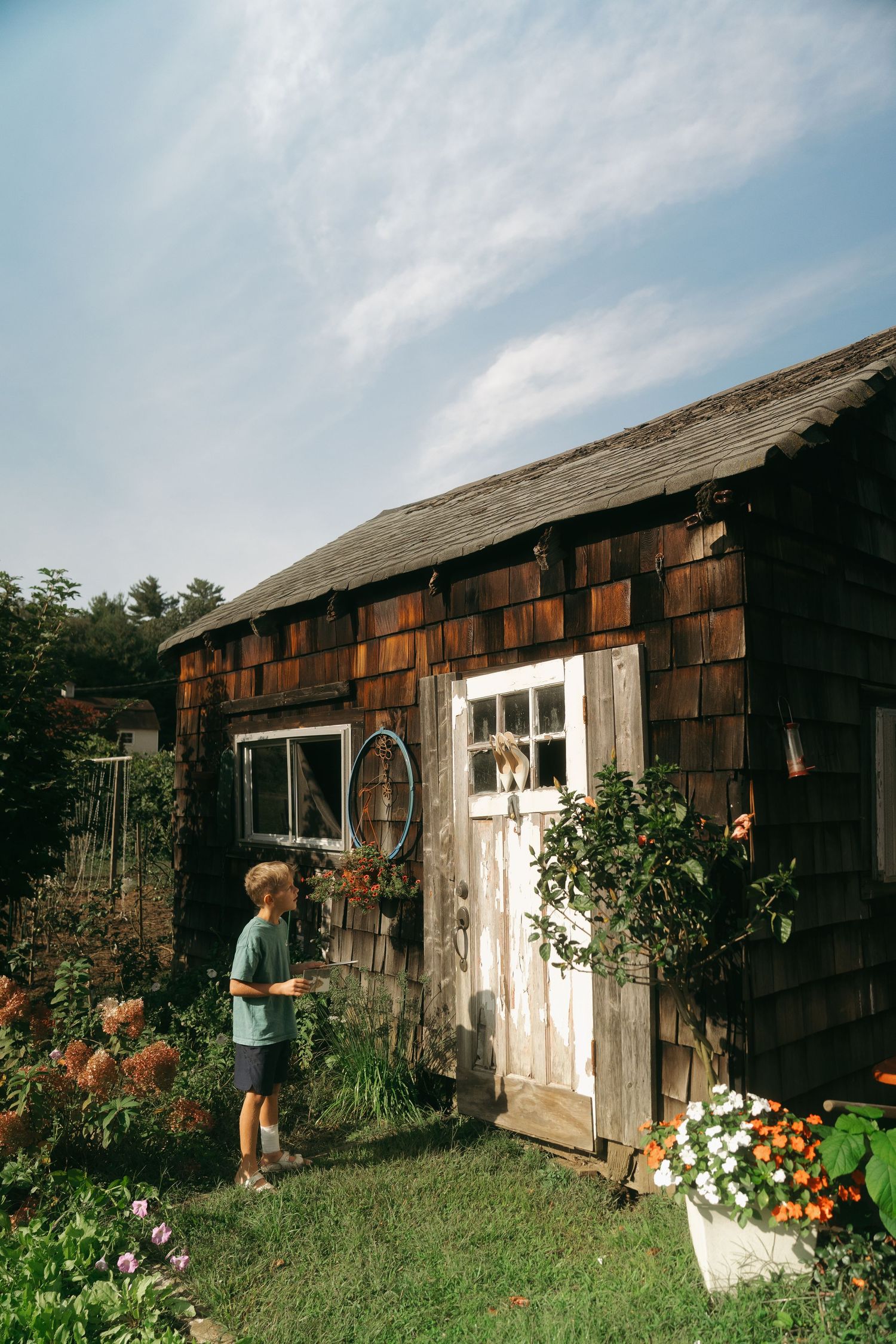 A rustic wooden garden shed with cedar shingles surrounded by colorful flowers and greenery on a summer day.