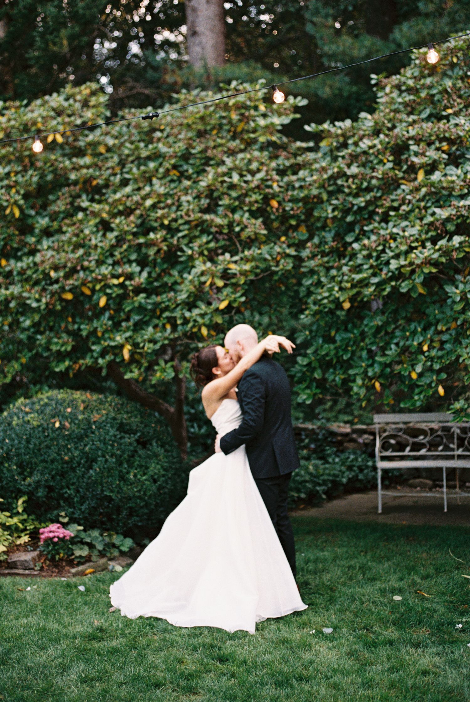 Wedding couple dances and twirls on a manicured lawn surrounded by garden foliage.