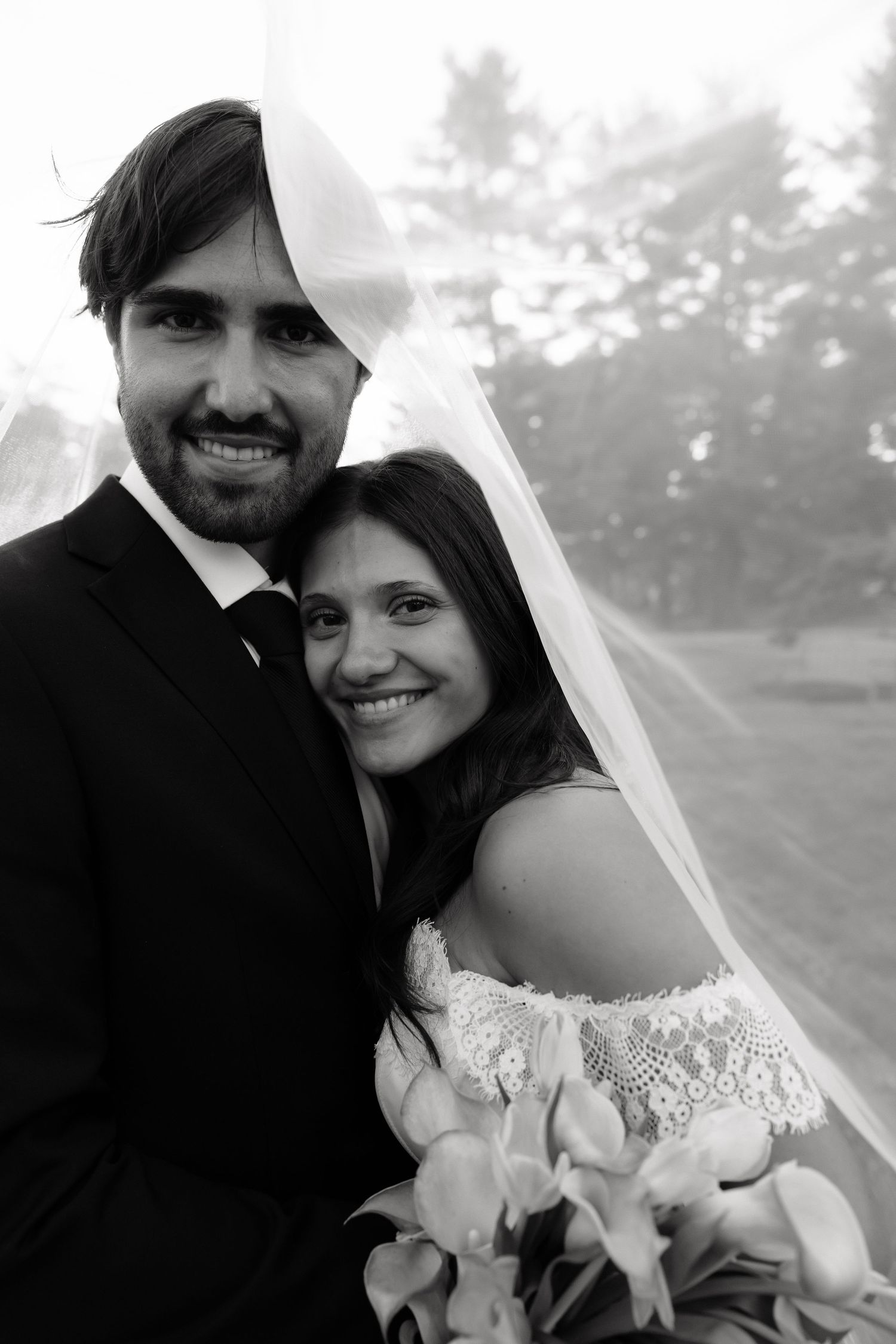 Black and white portrait of smiling bride and groom under wedding veil outdoors.