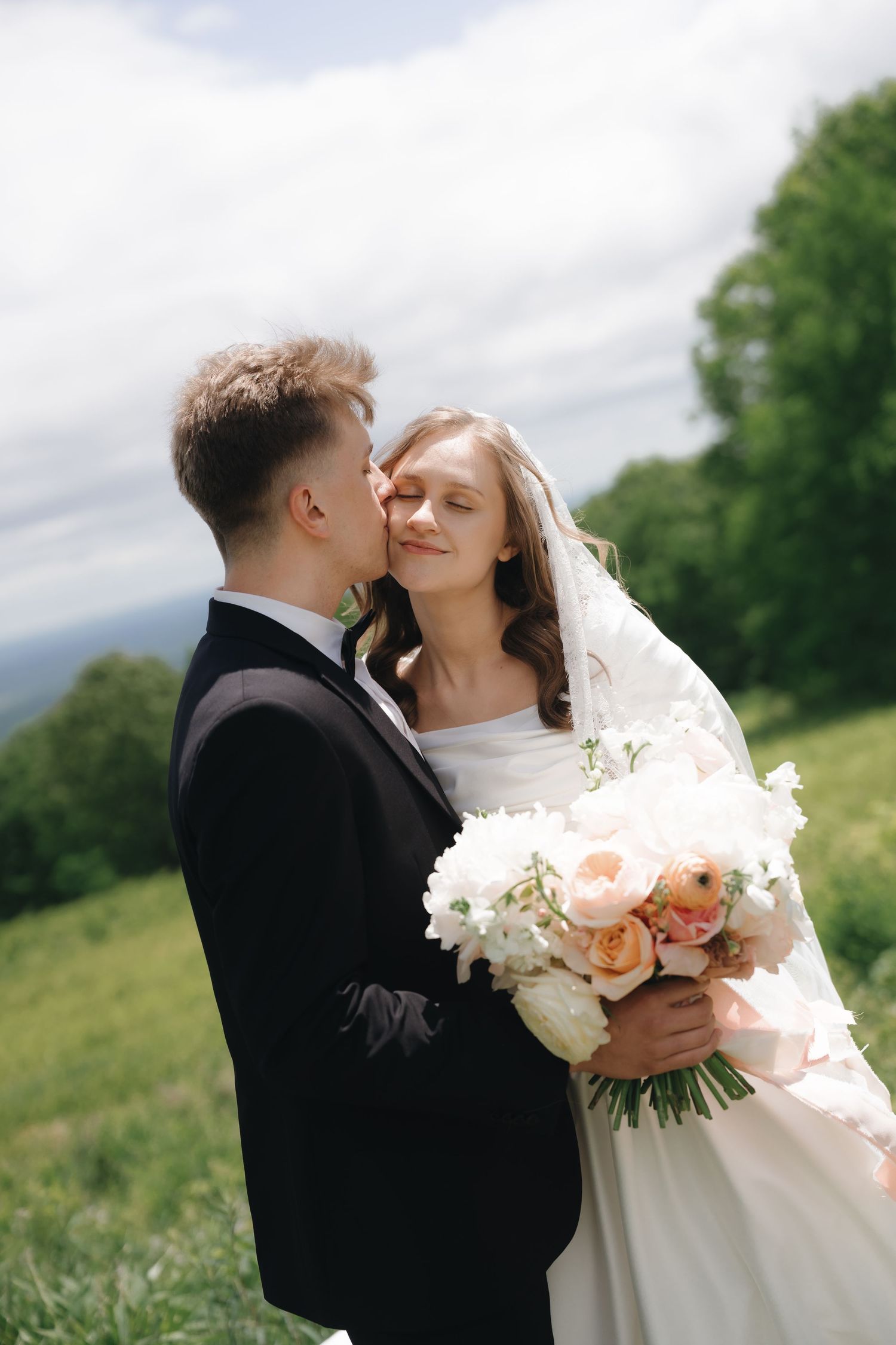 A couple shares an intimate moment in a meadow while holding a beautiful bouquet of pink and white flowers.