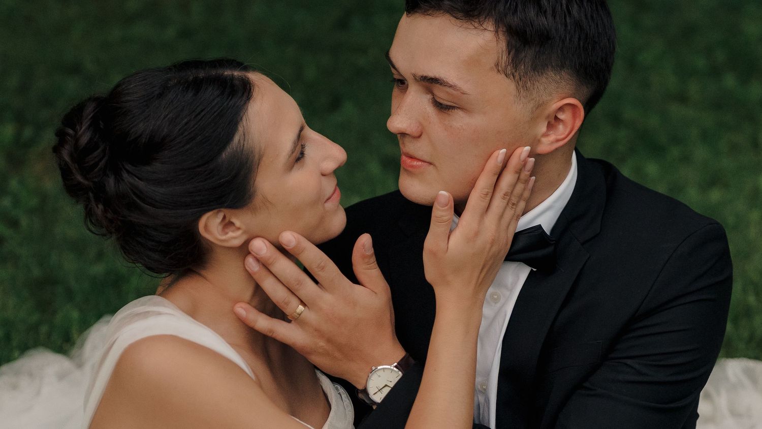A bride and groom tenderly hold each other's faces in an intimate wedding portrait outdoors.