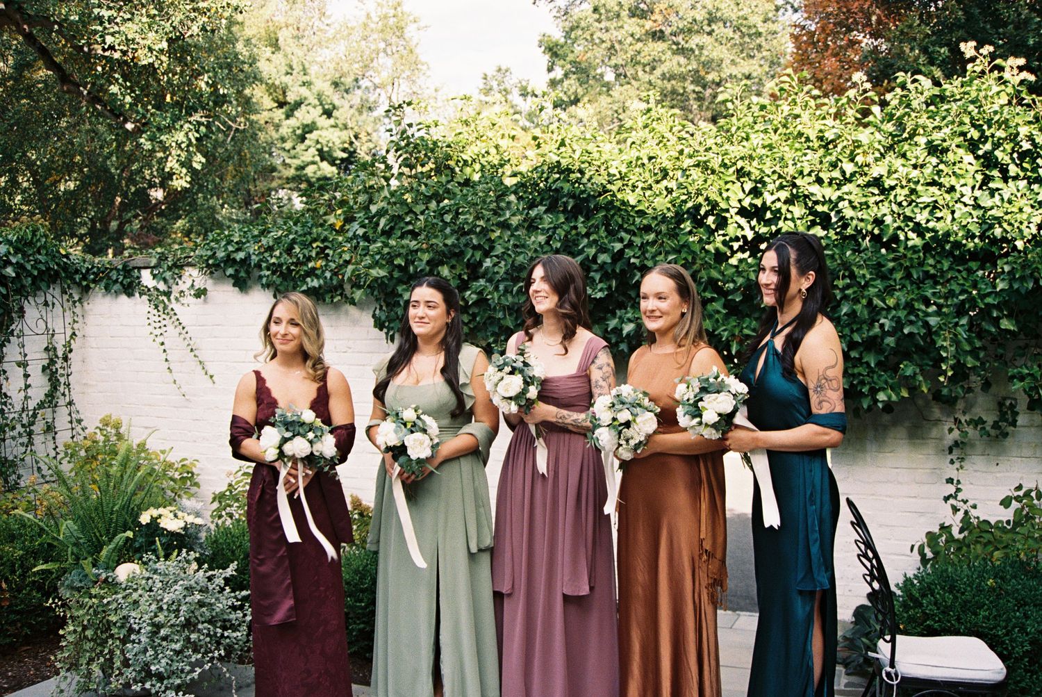 Wedding party members in coordinating dresses of burgundy and sage green standing in a garden.