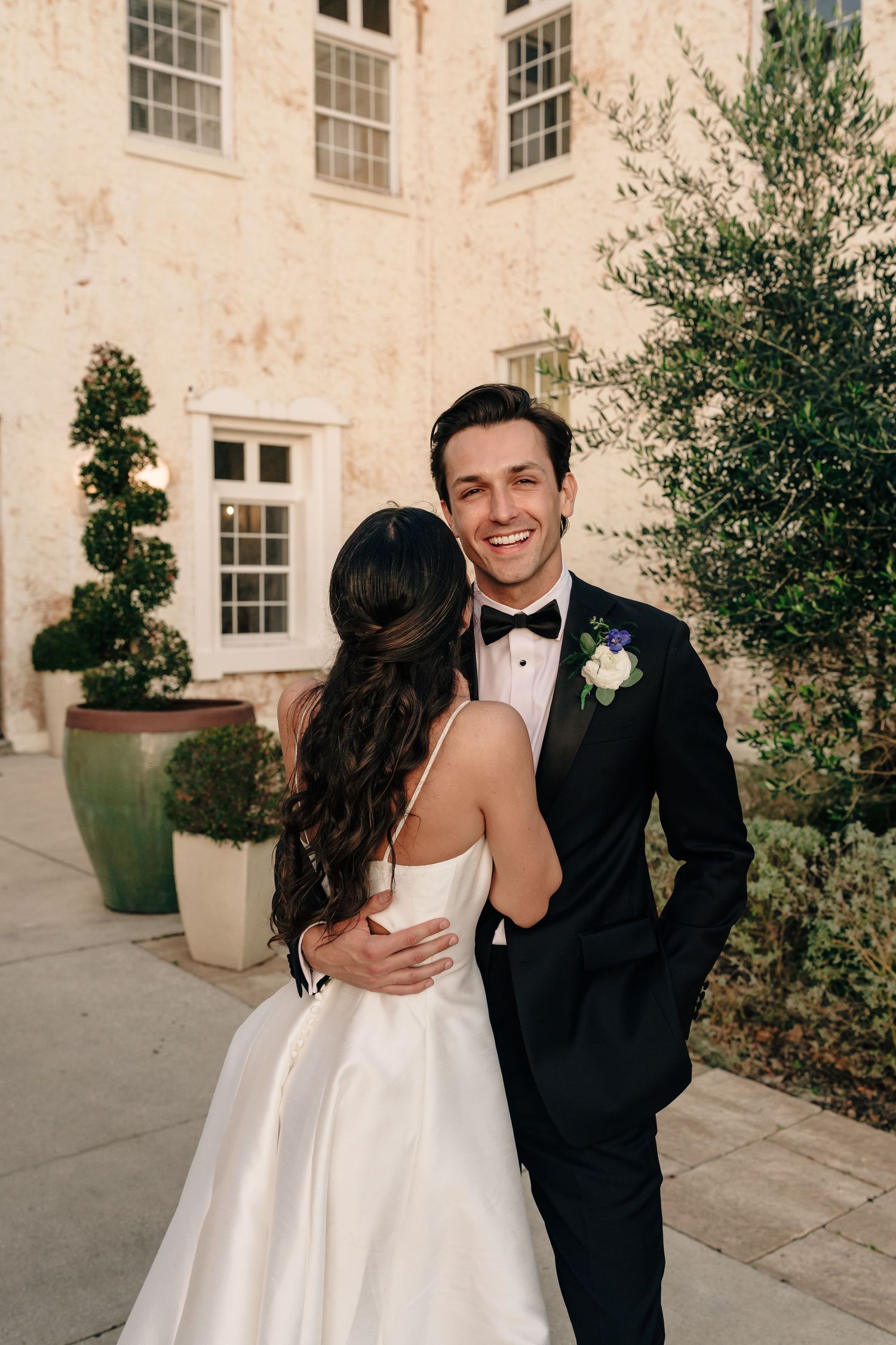 A bride and groom share a joyful moment together outside a building with white walls and elegant windows.