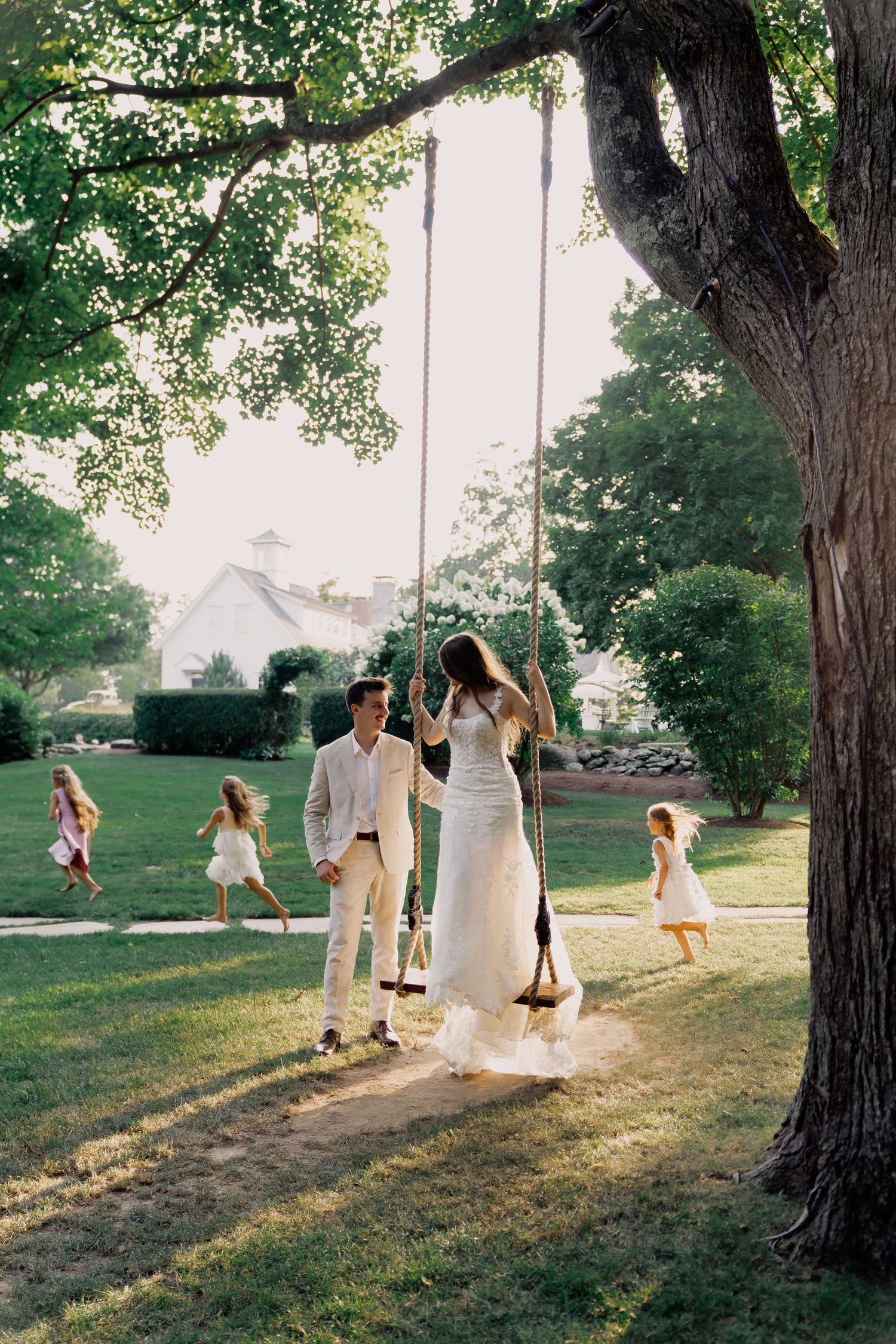 A couple shares a romantic moment on a tree swing in a sunlit garden while children play in the background.