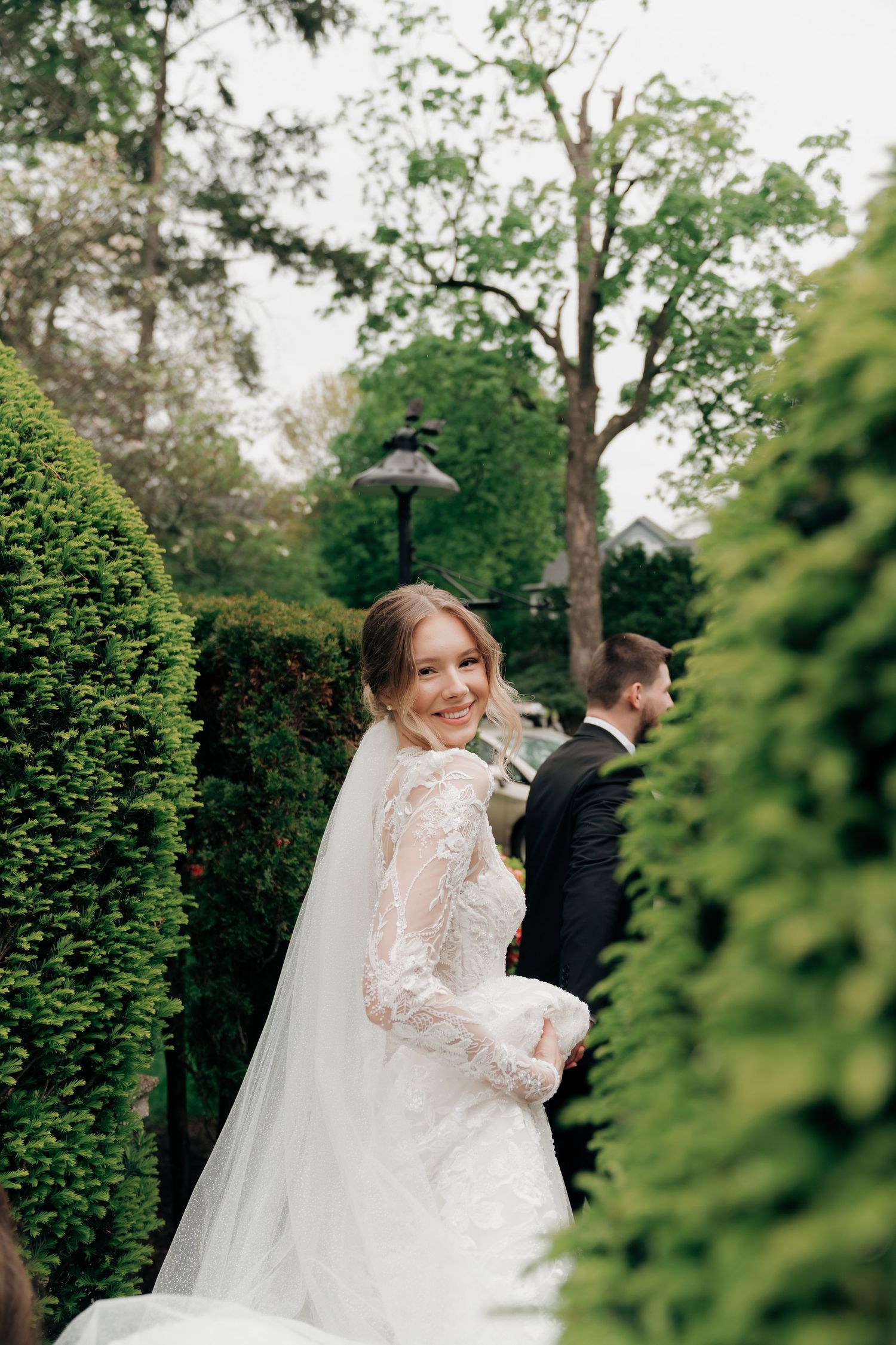 Smiling bride in lace wedding gown and veil stands in lush garden path with groom in background.