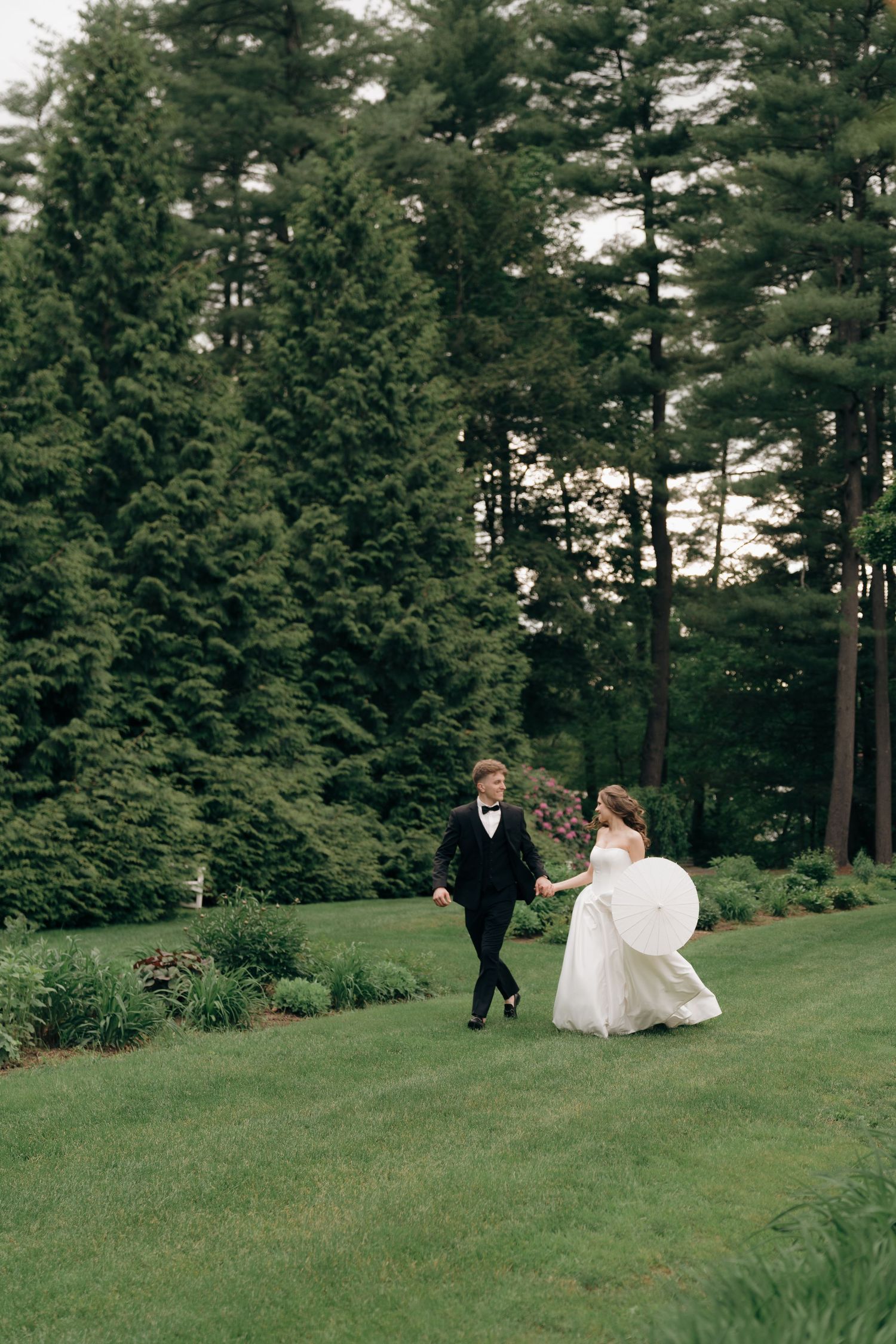 A wedding couple walks through a scenic pine forest setting on a grassy hillside.