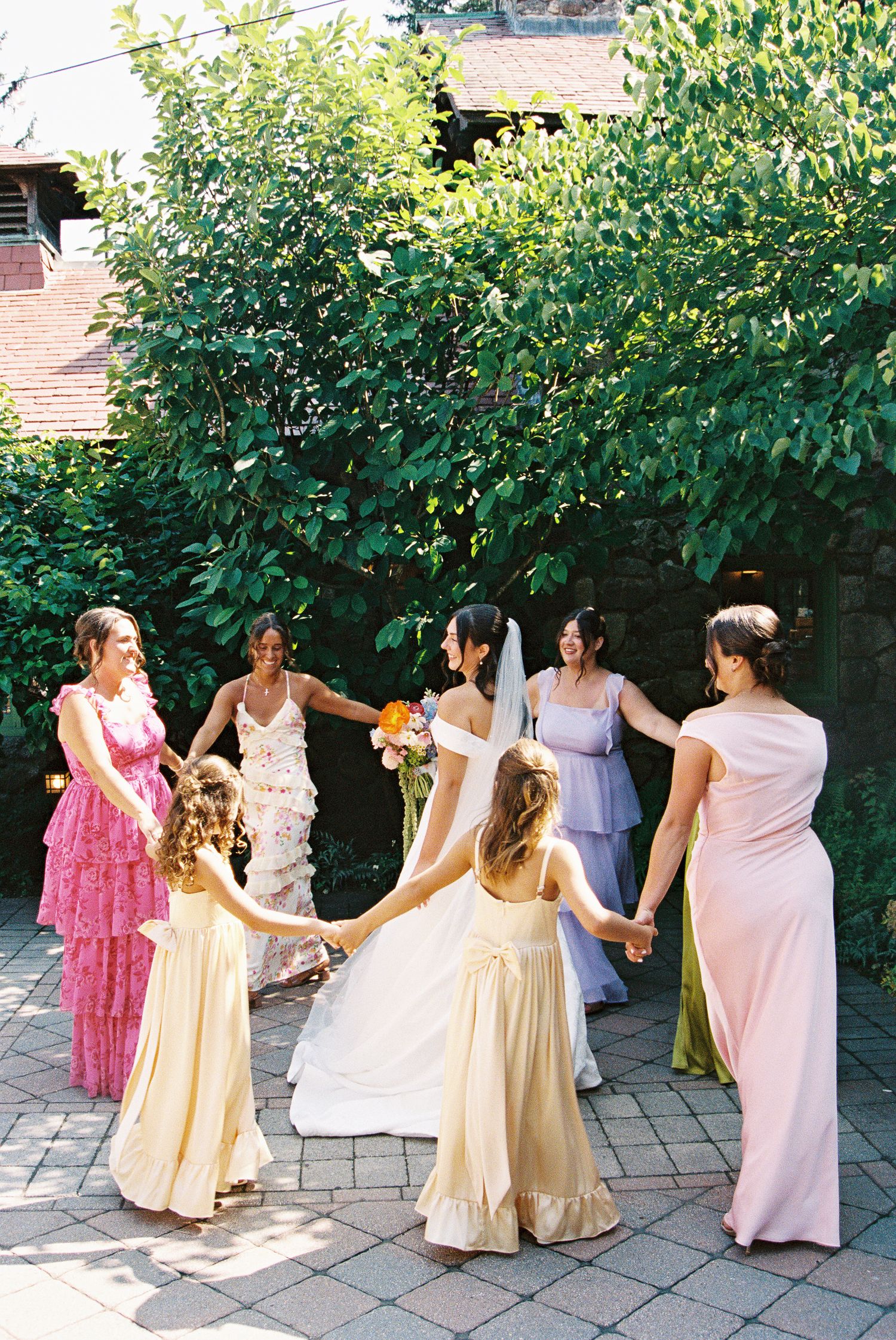 Bride and bridesmaids in colorful gowns hold hands in a circle outdoors, celebrating a joyful wedding day.