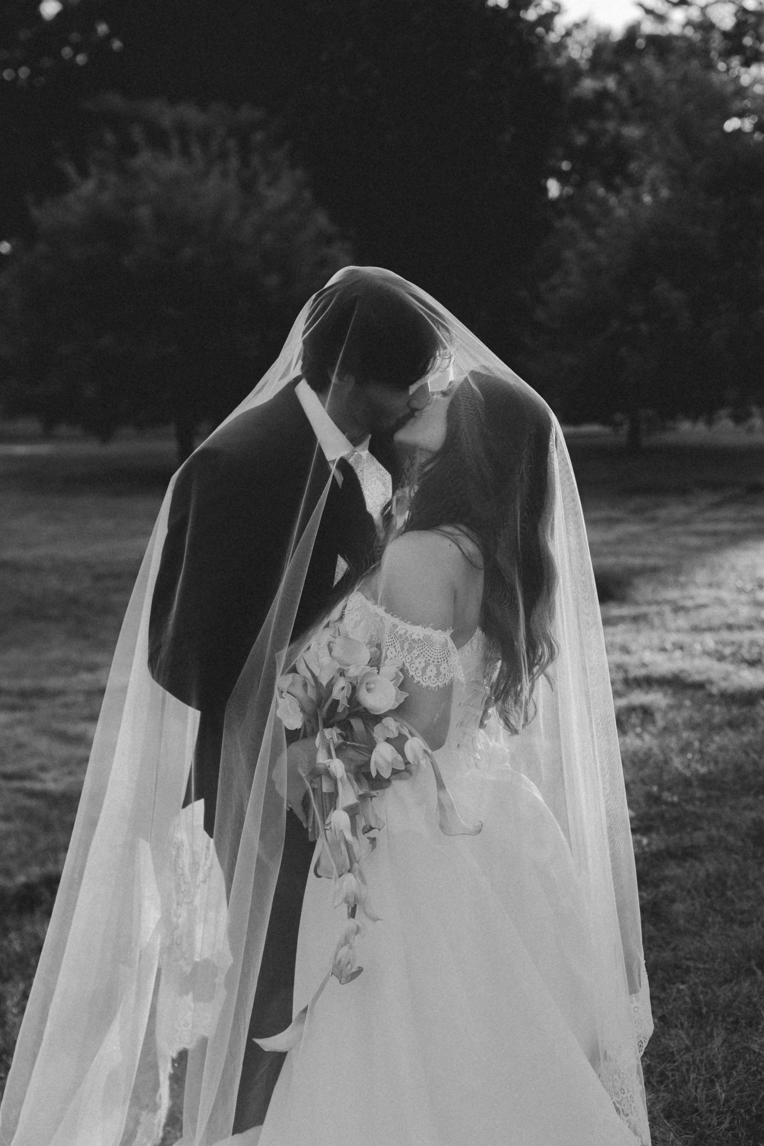 Black and white photo of bride and groom kissing under wedding veil outdoors.