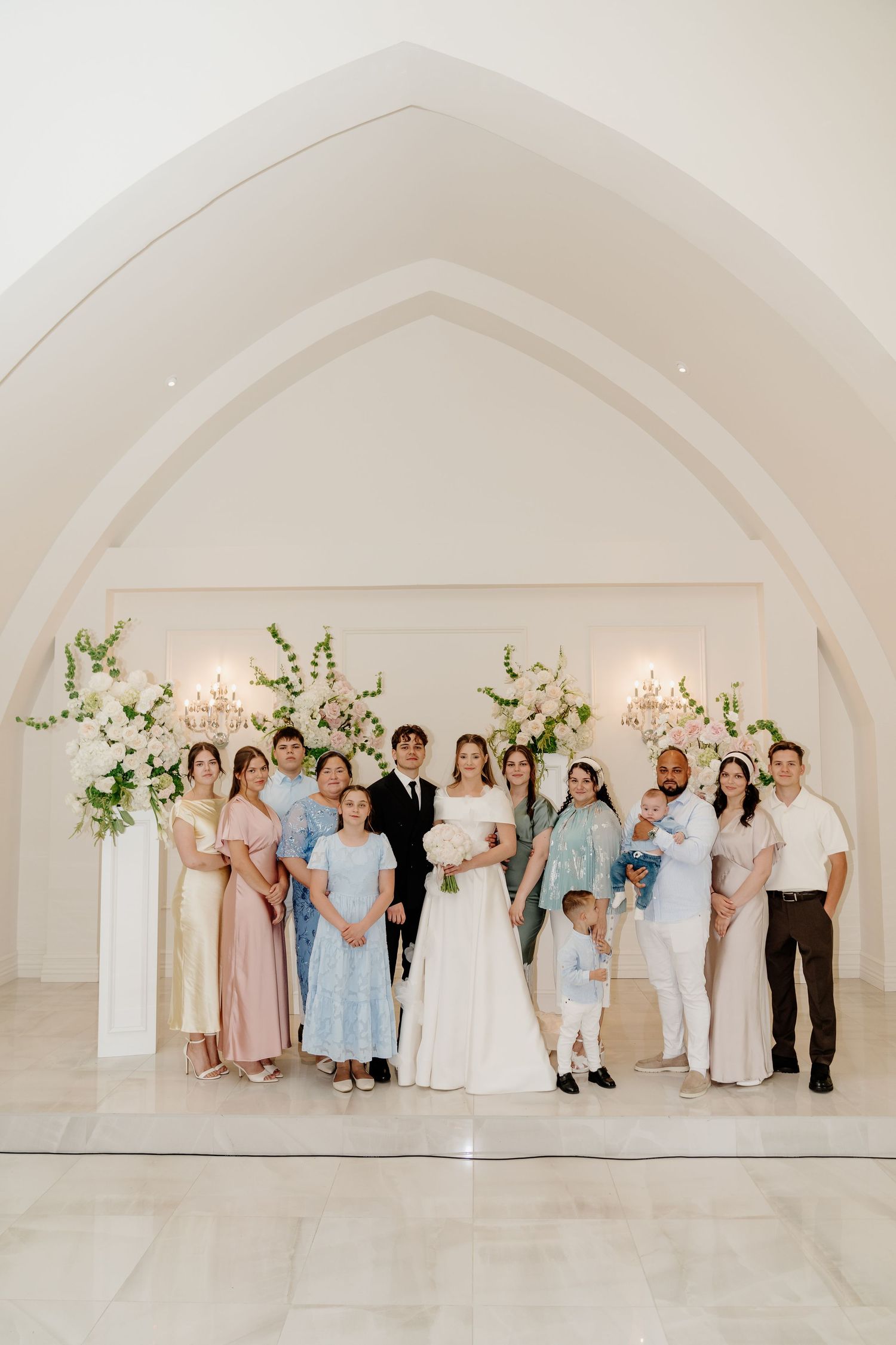 Wedding party poses under white arched altar with floral arrangements in elegant bright venue.