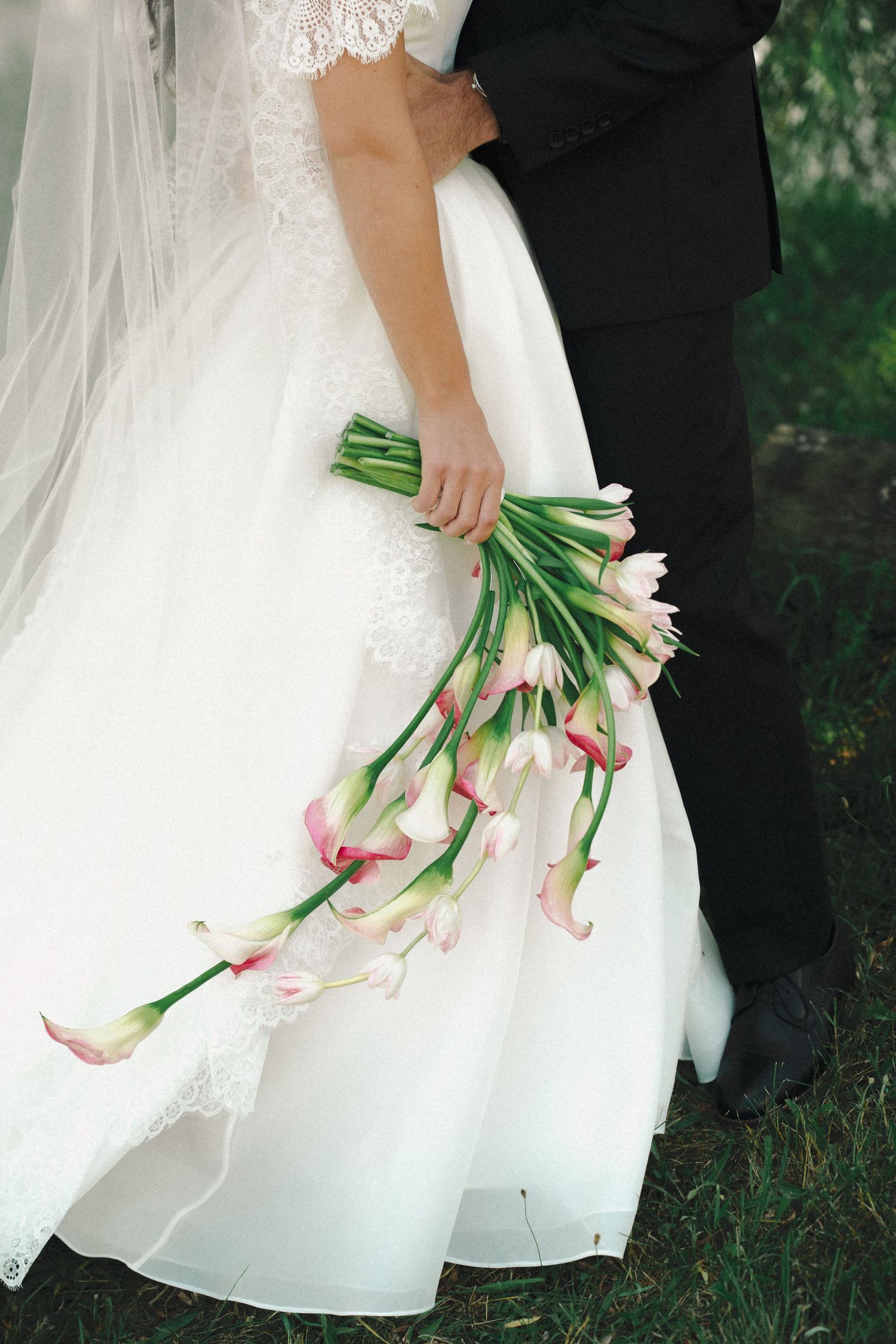 Bride in white lace gown holding cascading pink calla lily bouquet while standing with groom in black suit outdoors.
