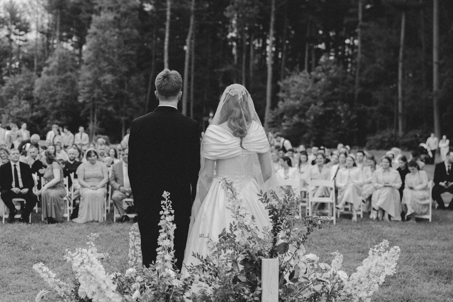 Black and white photo of wedding guests seated in rows during an outdoor ceremony with trees in background.
