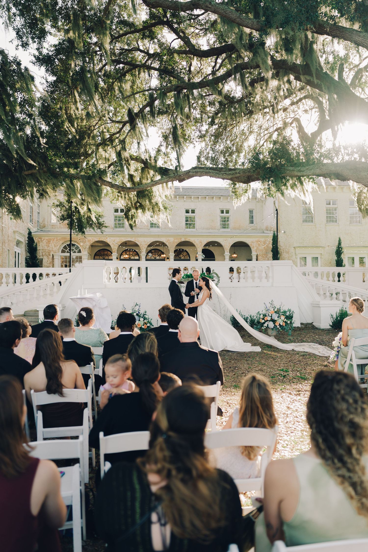 Outdoor wedding ceremony setup with white chairs and arches under moss-draped oak trees.